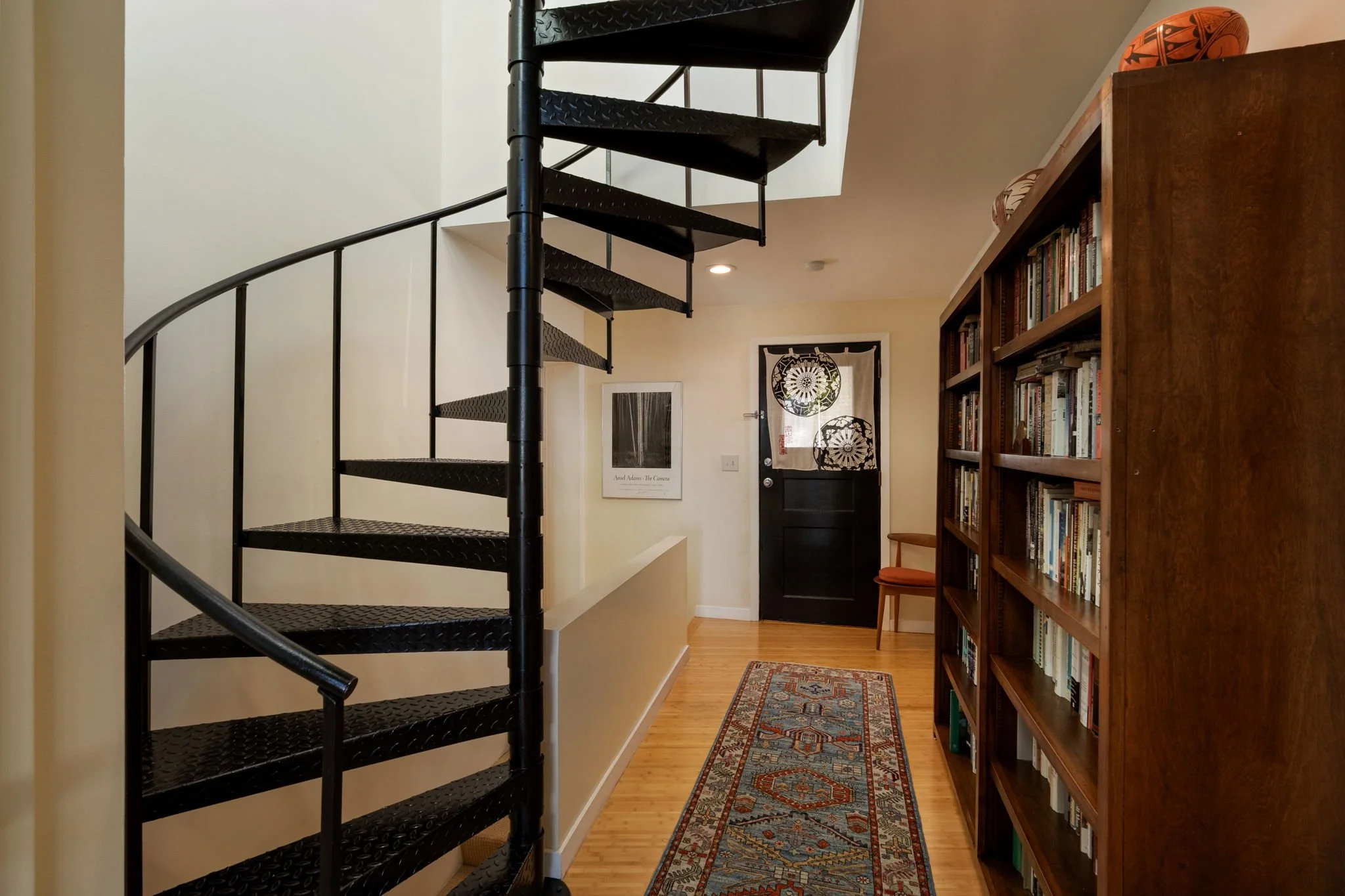 Interior view of a home featuring a black metal spiral staircase on the left, a bookshelf filled with books on the right, a door in the background with a decorative fabric curtain, a small chair next to the bookshelf, and a colorful oriental rug on t