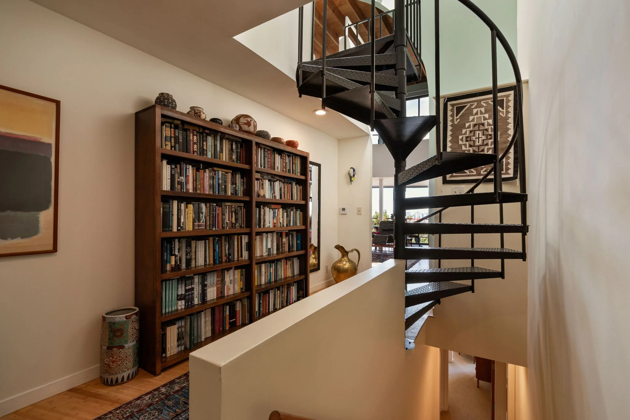 Interior view of a modern home featuring a spiral staircase with black metal steps, a tall wooden bookshelf filled with books and decorative pottery, a abstract painting on the wall, a large window allowing natural light, and an intricately patterned