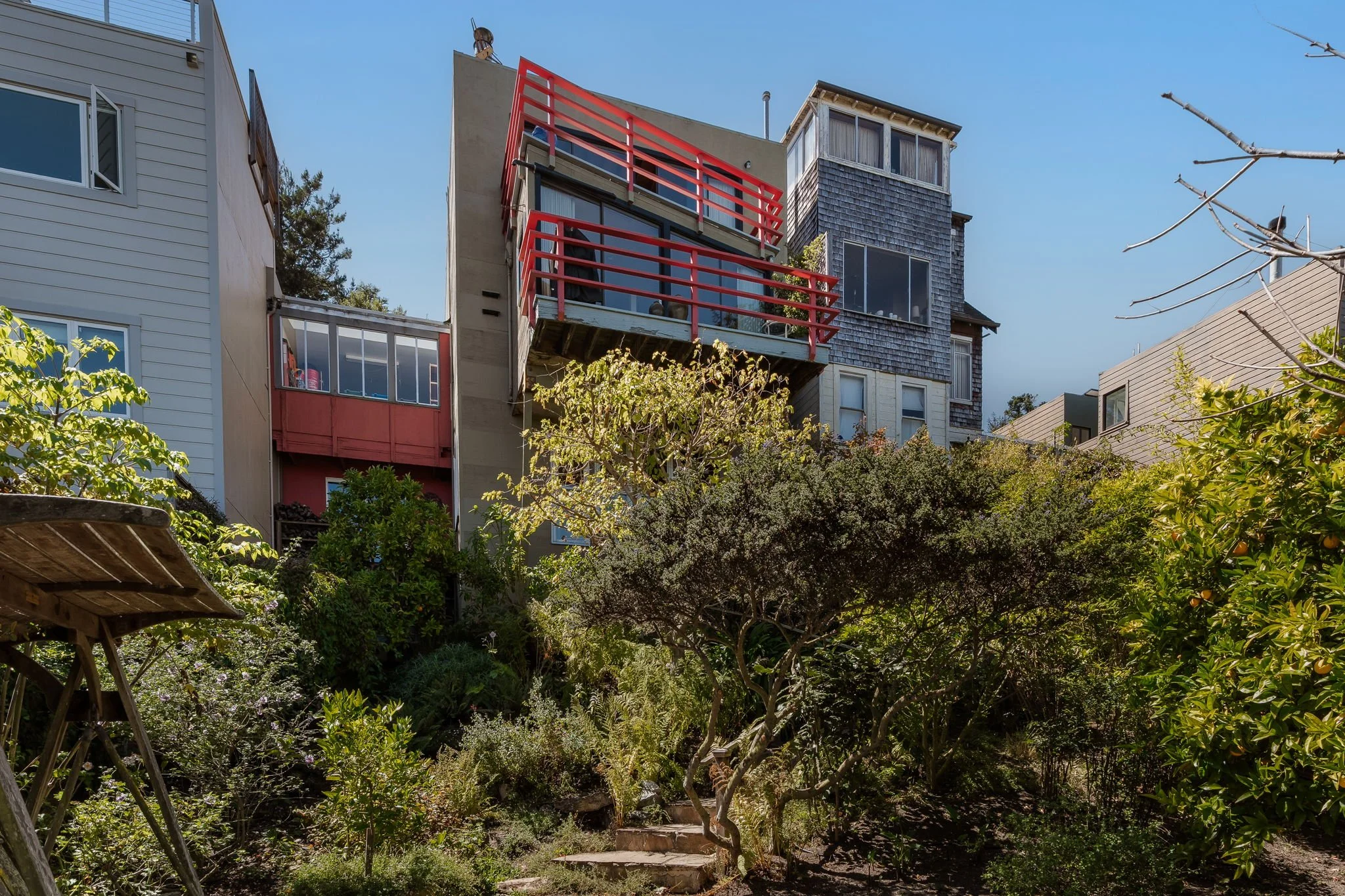 Multiple neighboring houses with various architectural styles and colors are seen in a hillside neighborhood, with lush garden in the foreground and a clear blue sky above.