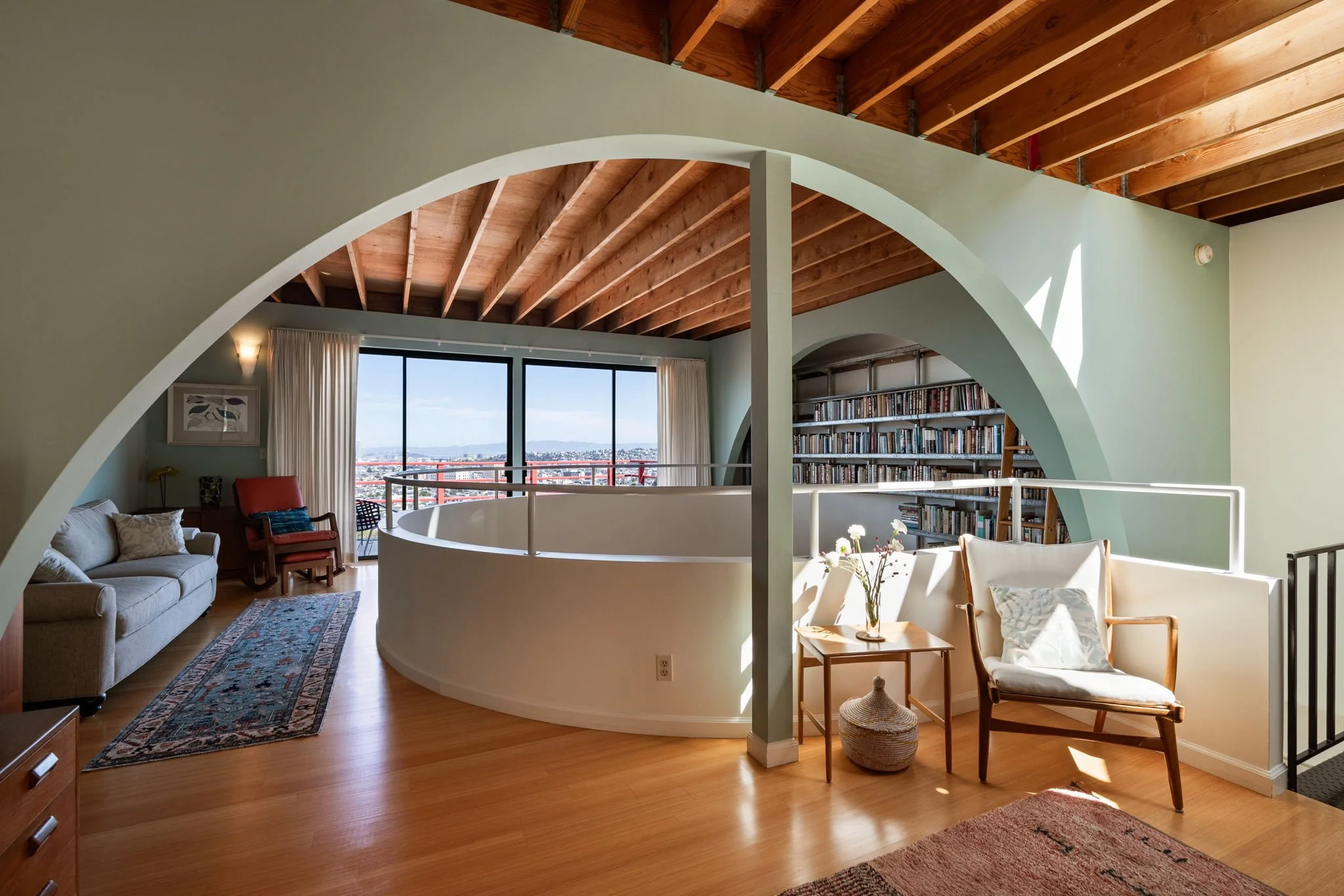 A spacious living room with hardwood floors, a curved white wall with a railing, a large window with a city view, a patterned rug, beige sofa, red armchair, bookshelves, and armchairs with cushions, and wooden ceiling beams.