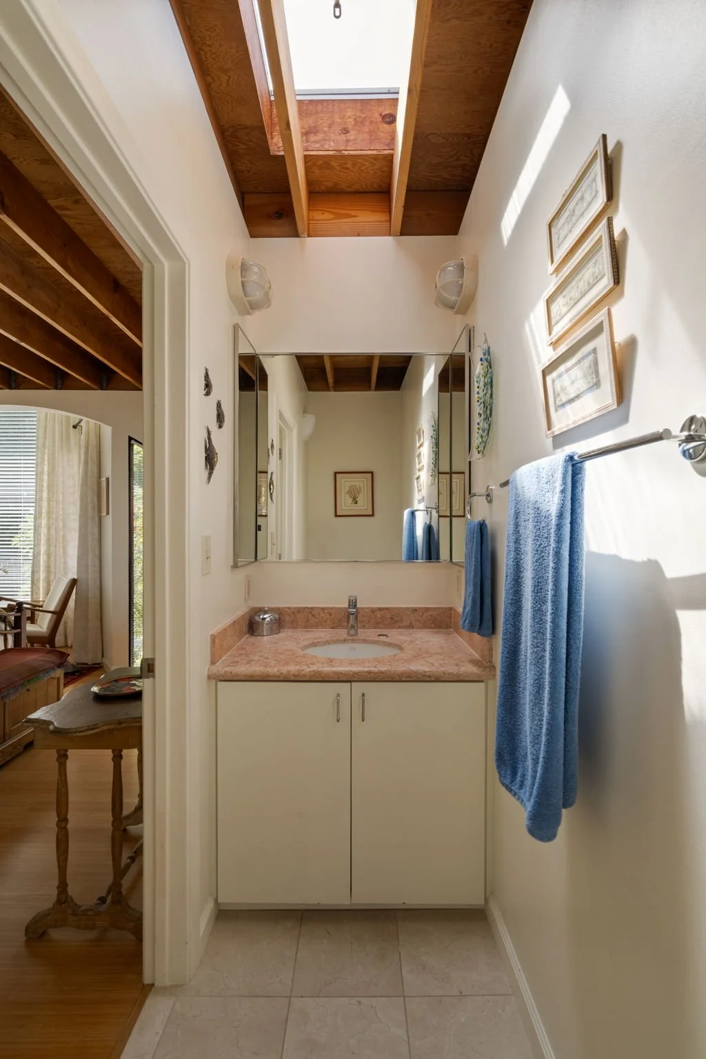 Bathroom with a pink marble countertop sink, a large mirror, blue towels hanging on a silver towel rack, framed artwork on the wall, and a skylight showing wooden ceiling beams.