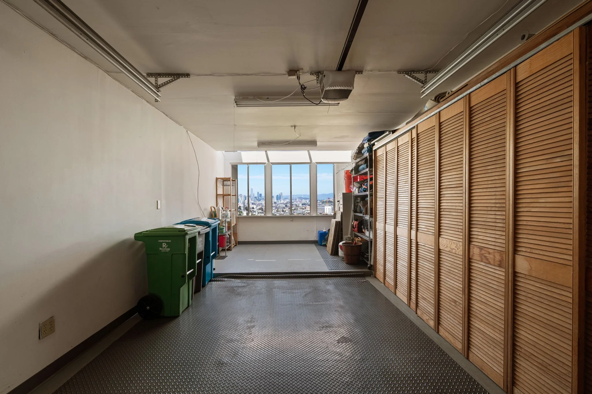 Empty laundry or storage room with large window showing city skyline, green and blue recycling bins, wooden cabinets, and shelves with cleaning supplies.