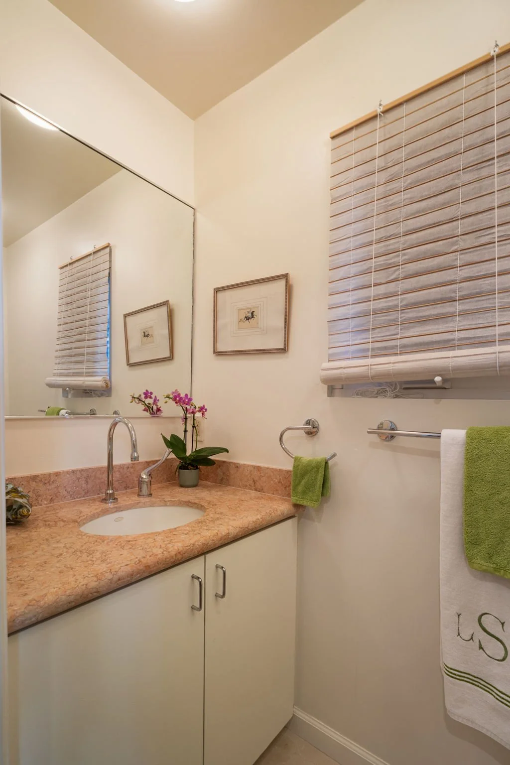 Bathroom with beige countertop, a built-in sink, a mirror, framed artwork, a potted orchid, towel bars holding green and white towels, and a window with wooden blinds.