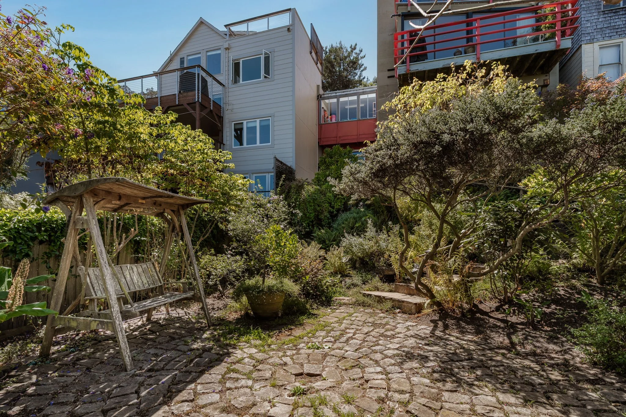 A backyard garden with a wooden bench swing, lush green trees and shrubs, a cobblestone pathway, and multi-story houses with balconies and windows in the background.