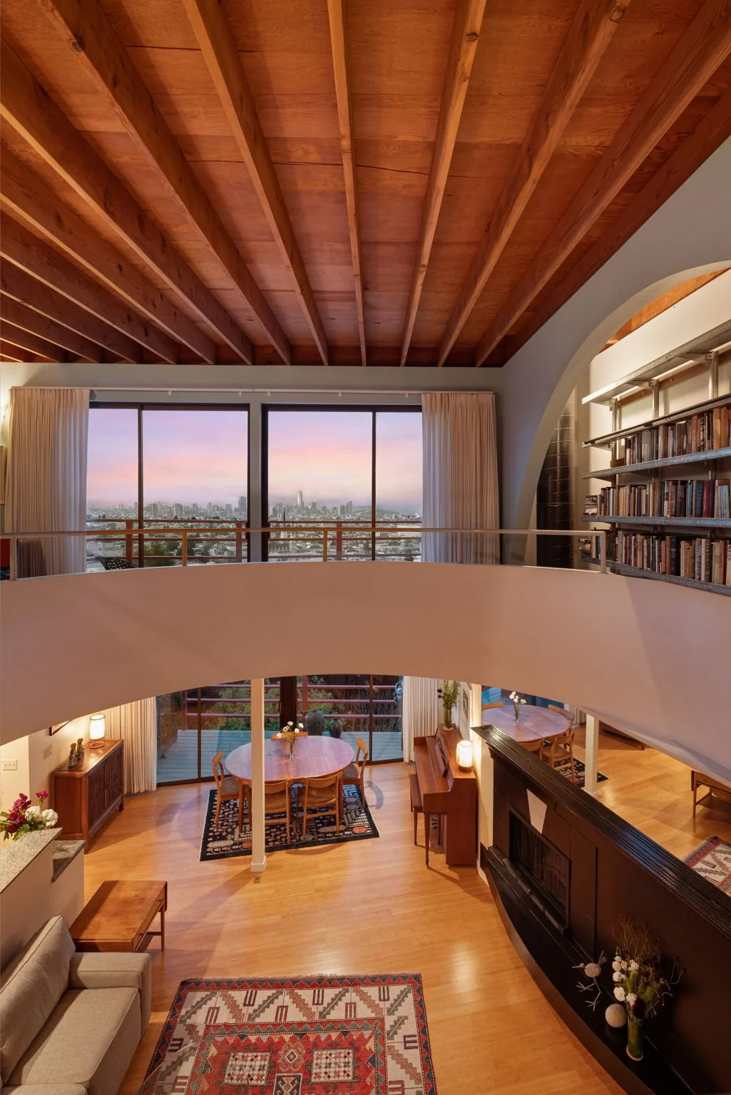 A spacious living room with a wooden ceiling and large windows showing a city skyline at sunset. The room features a round dining table, a bookshelf, a sofa, and a fireplace with decorative items.
