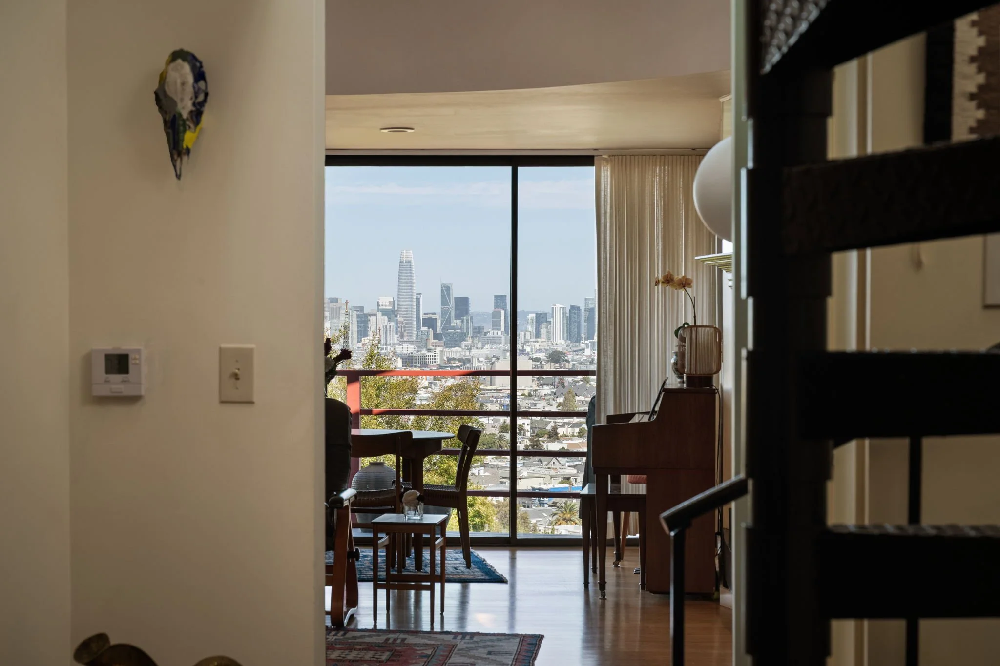 Living room with large window showing city skyline, furniture including chairs and a table, and a potted plant.