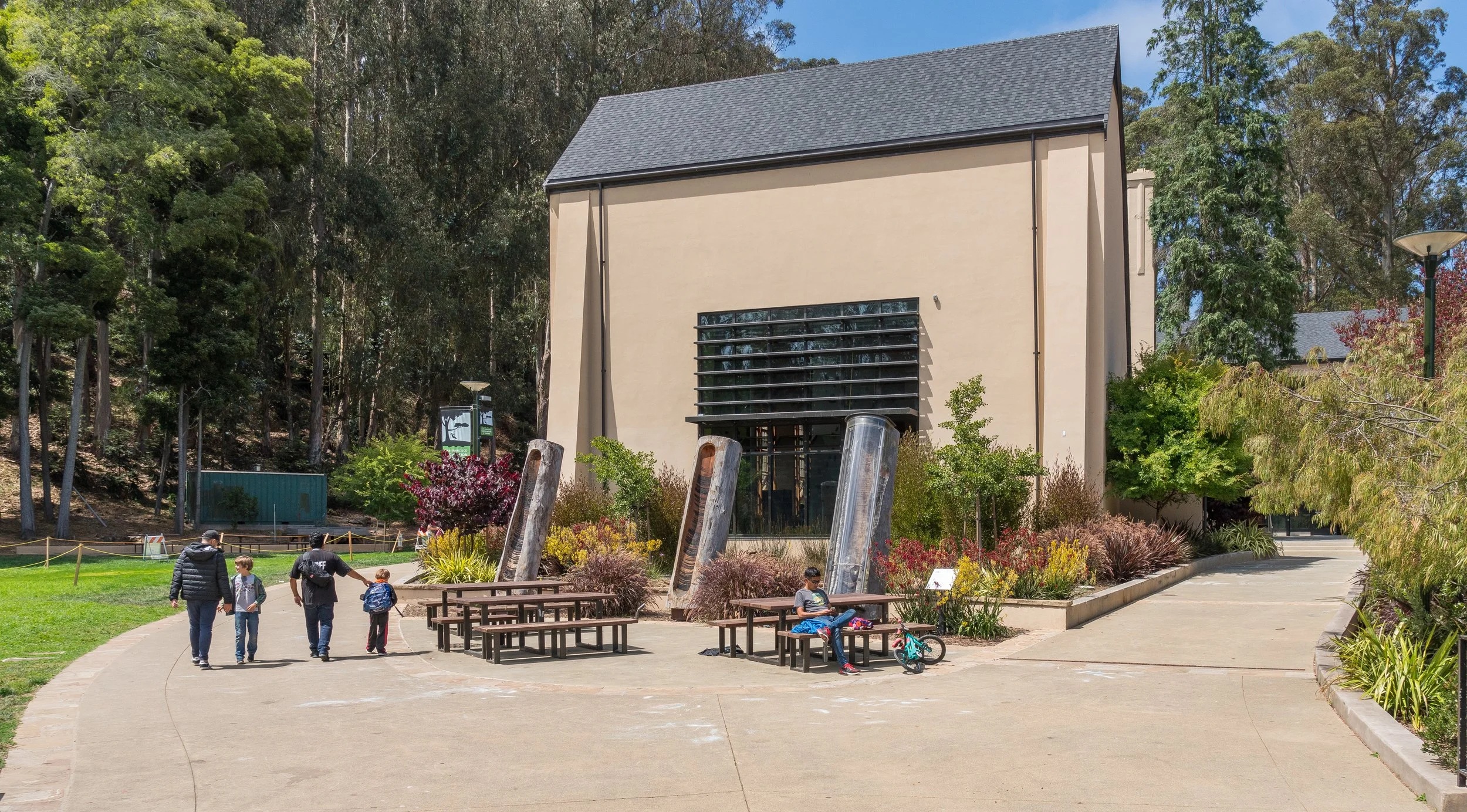 People walking and sitting in a park with benches, surrounded by trees and colorful plants, in front of a modern building with large windows and a gray roof.