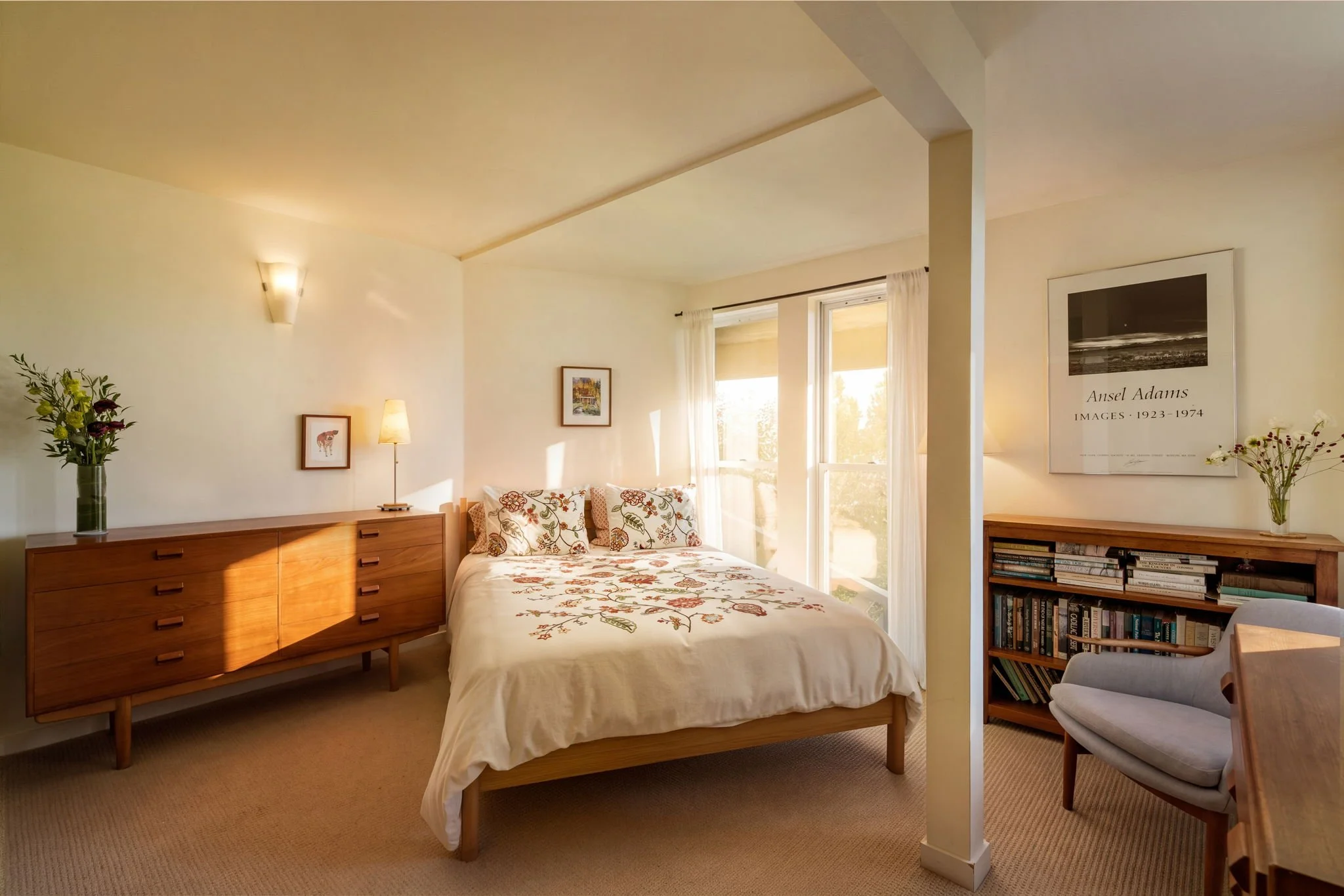 A bedroom with a bed featuring embroidered floral bedding, a wooden dresser, framed artwork on the walls, a window with sheers, a table with books, and a gray upholstered chair.