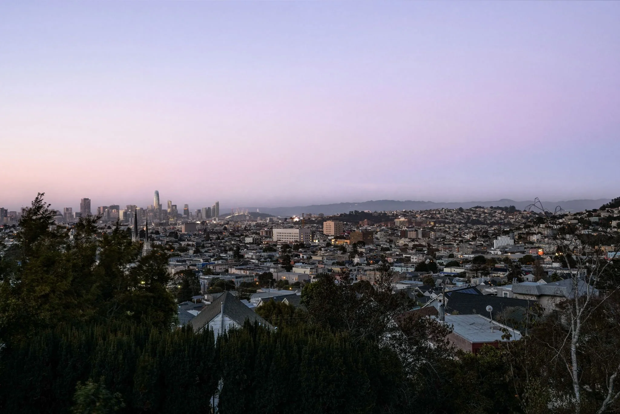 Panoramic view of a city skyline at dusk with a large building under construction, surrounded by residential and commercial buildings, with hills in the distance and a pastel-colored sky.