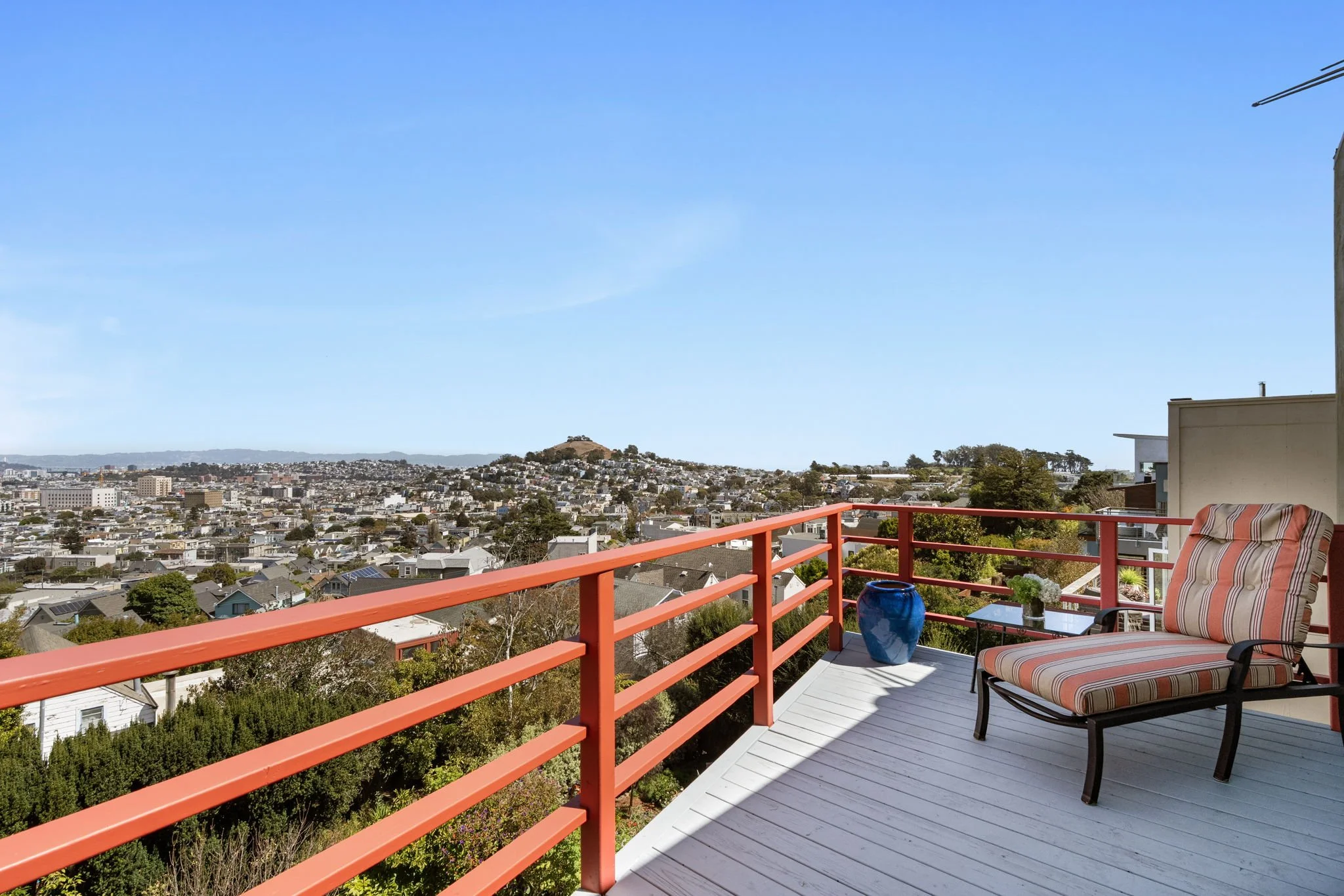 Balcony with a red railing, striped outdoor chair, blue vase, and view of a cityscape with houses, trees, and hills under a blue sky.