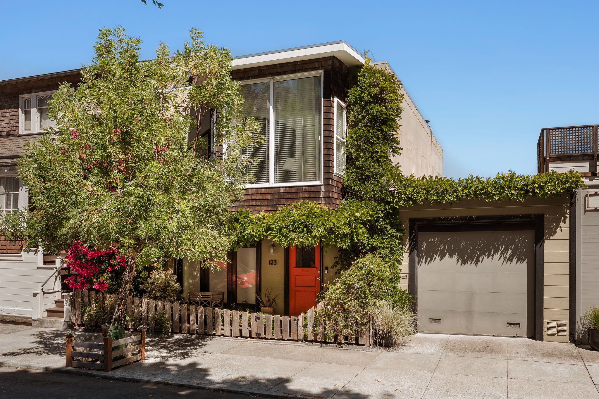 A modern residential house with a mix of wood and stucco exterior, a bright orange front door, large windows with blinds, a garage door, and a small garden with trees, shrubs, and a fence, under a clear blue sky.