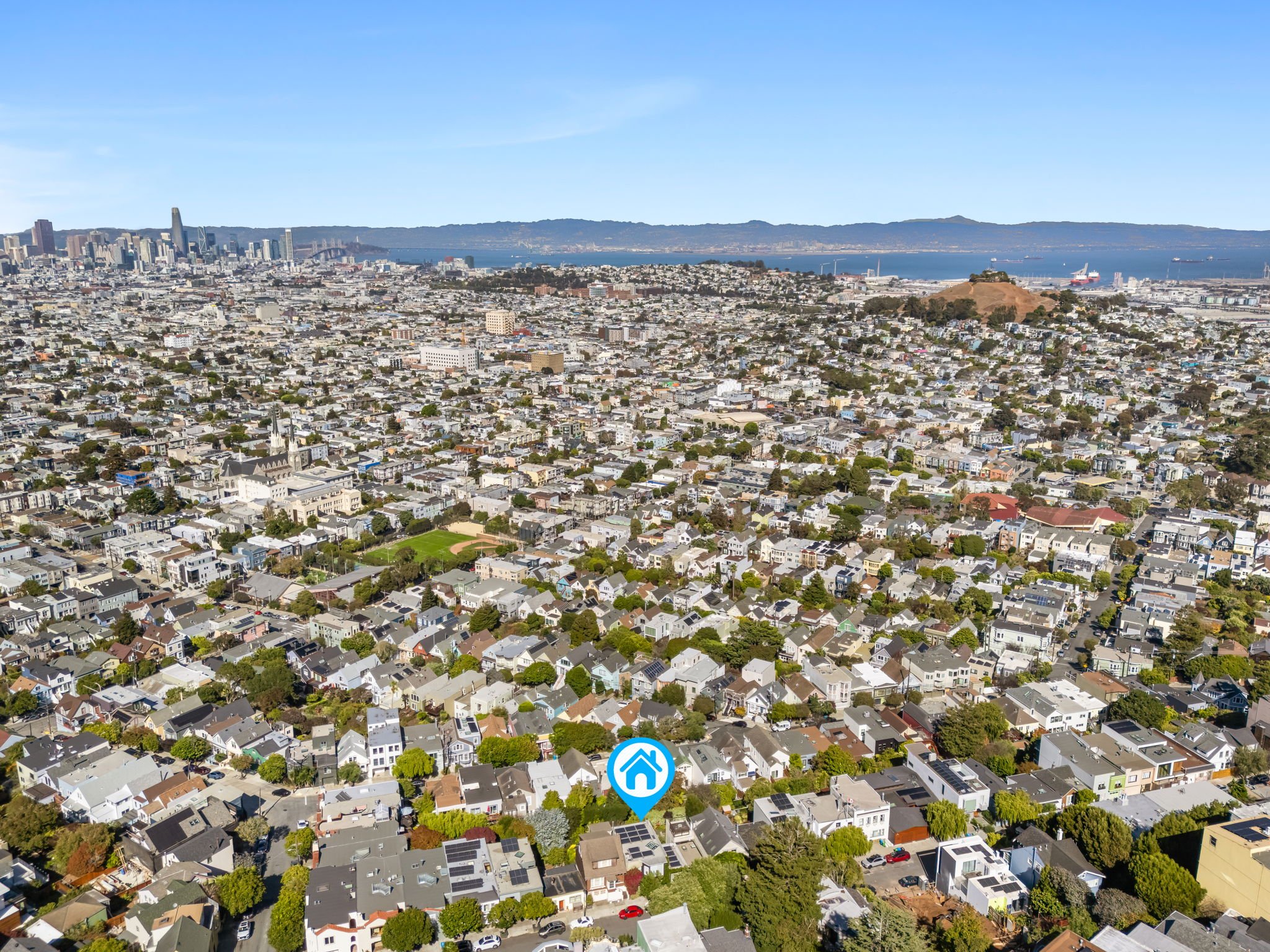 Aerial view of a densely populated coastal city with residential houses, a park, a hilltop with a structure, high-rise buildings, a body of water with ships, and a distant hilly coastline under a clear blue sky.