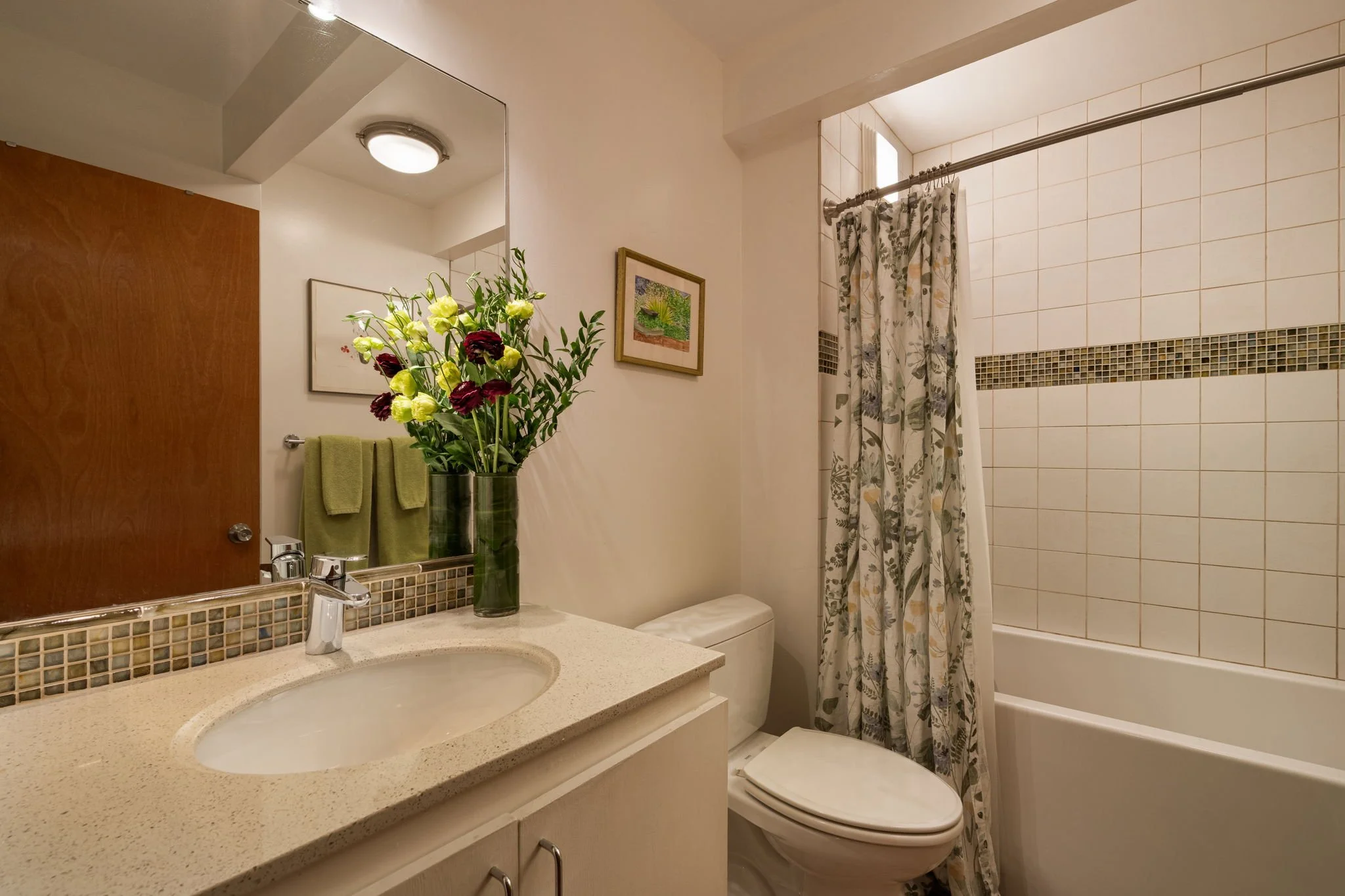 Bathroom with a vanity mirror, sink, green towels, a vase with flowers, framed pictures on the wall, a toilet, and a bathtub with a shower curtain.