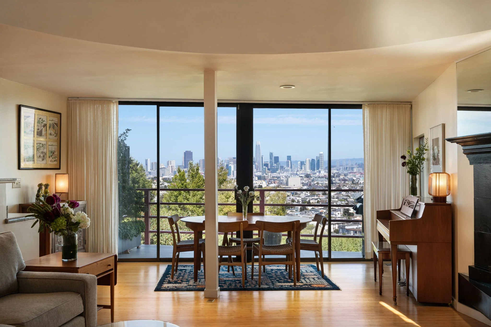 Living room with large windows showing city skyline, wooden dining table with chairs, piano, artwork, and houseplants.