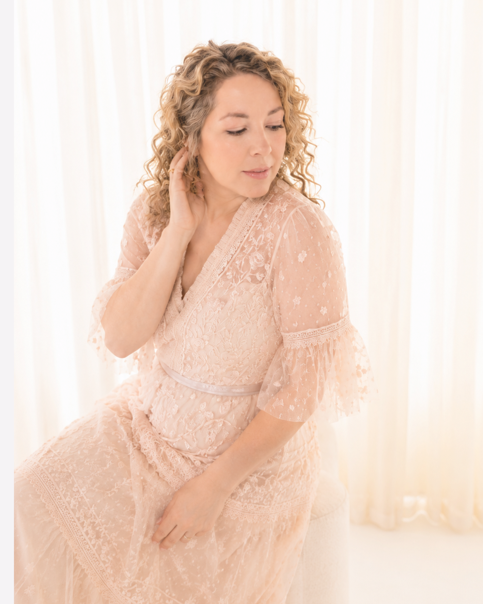 A woman in a light pink lace dress sitting by a window with sheer curtains, touching her hair and looking down.