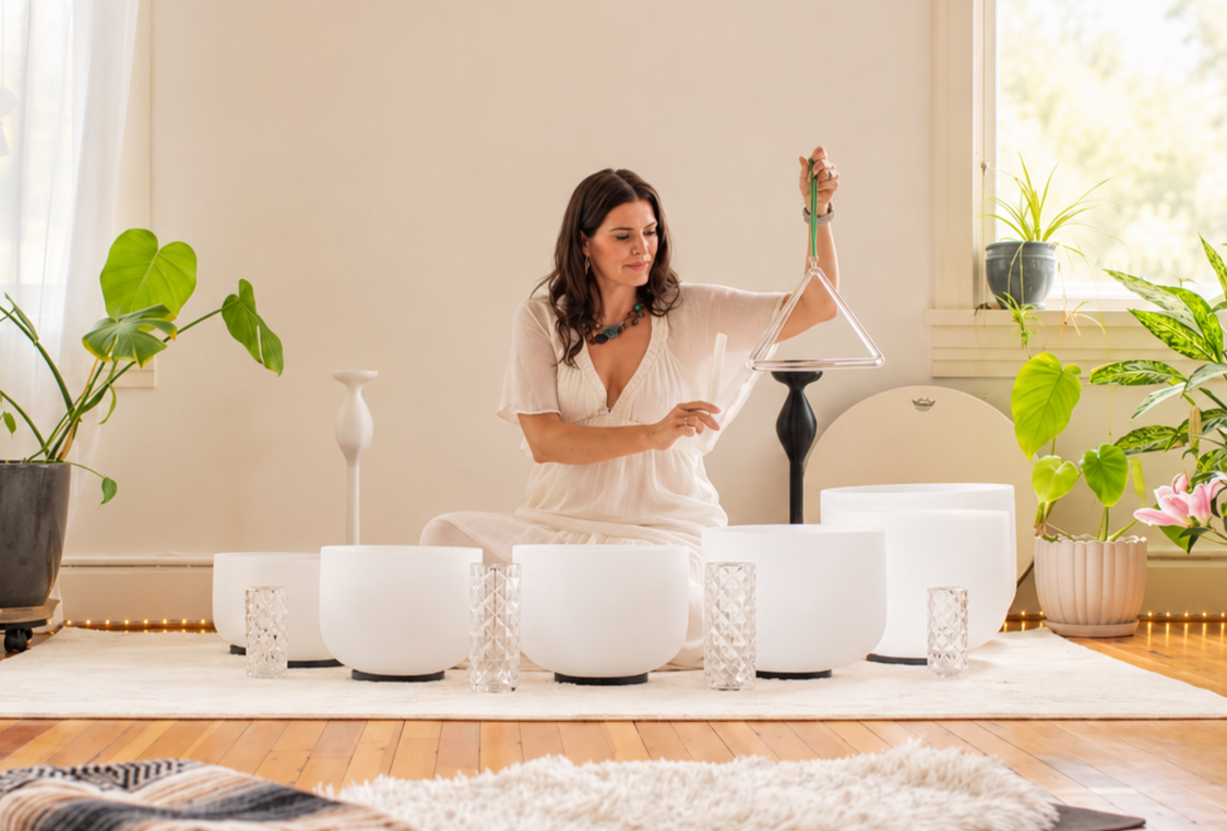 Woman sitting on rug in front of singing bowls and crystals, playing wind chimes, surrounded by plants.