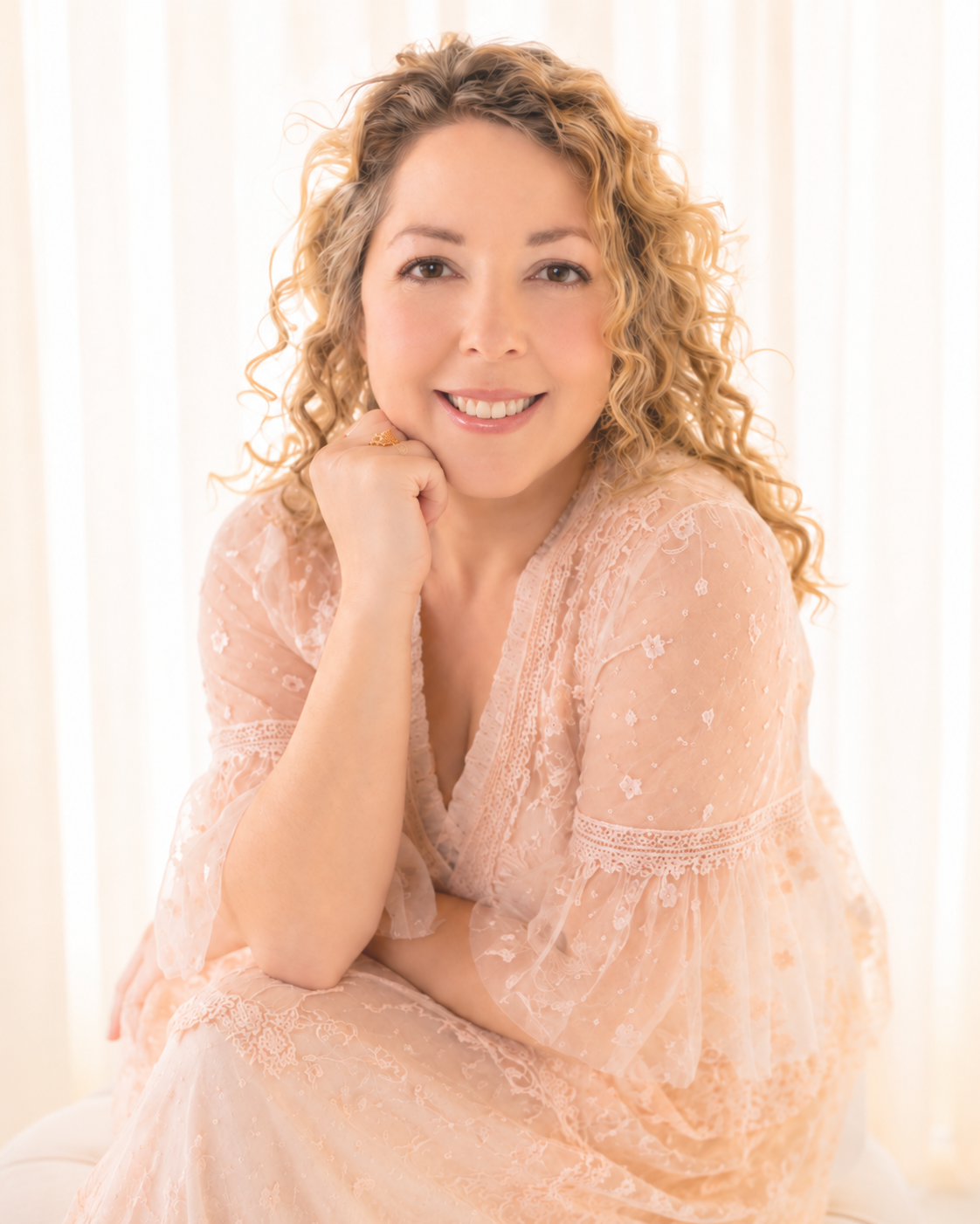 A woman with curly blonde hair smiling, resting her chin on her hand, wearing a peach-colored lace dress, in front of a light-colored curtain.