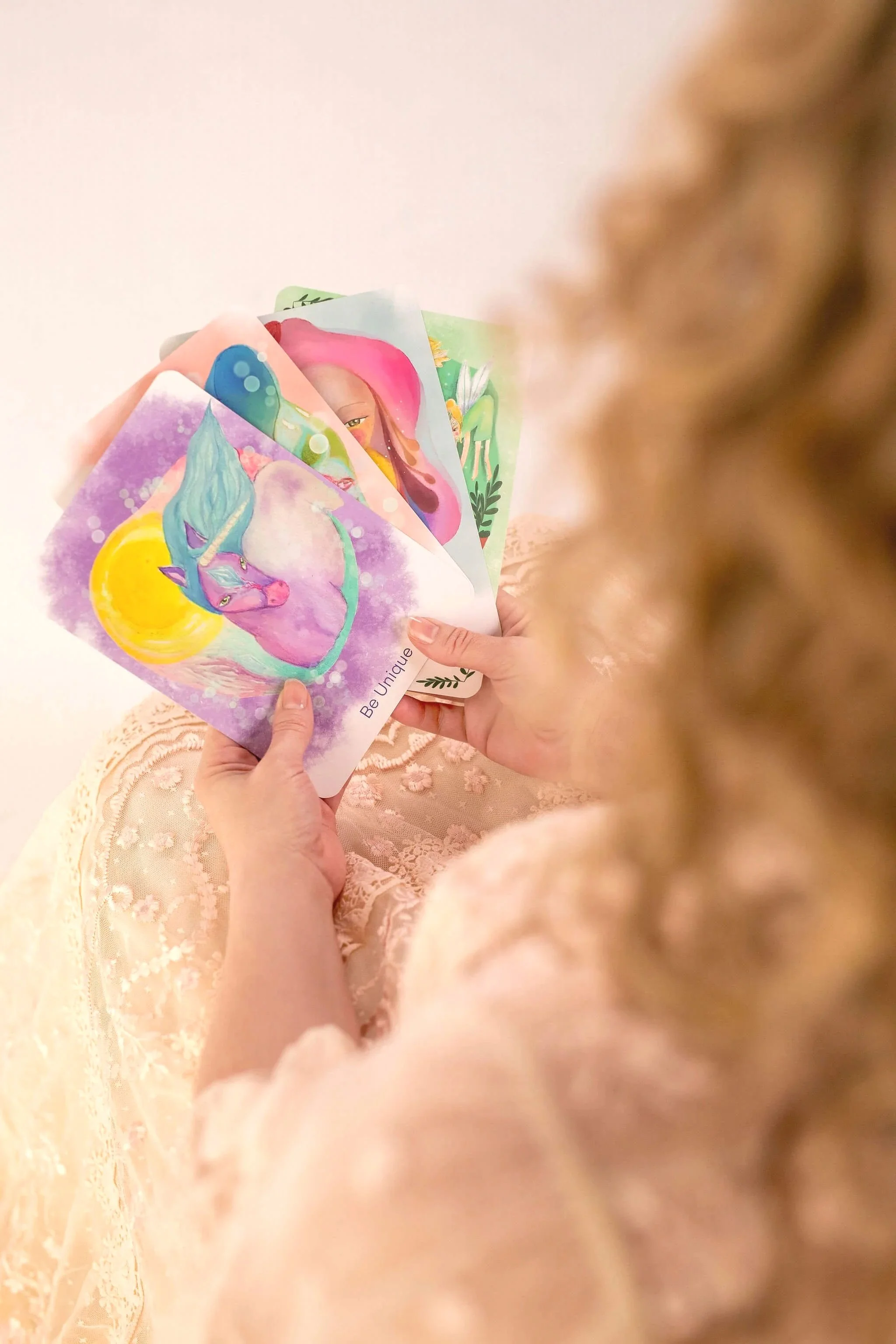 A person in lace clothing holding colorful illustrated cards with fantasy characters against a plain light background.