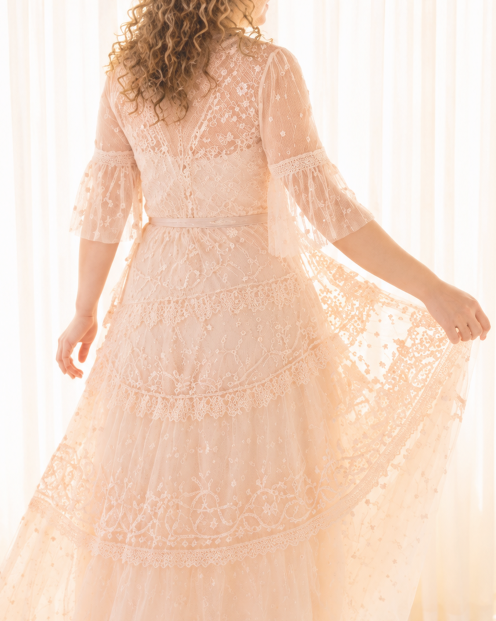 A woman wearing a peach-colored lace dress with intricate embroidery and sheer lace sleeves, standing in front of light-colored curtains.