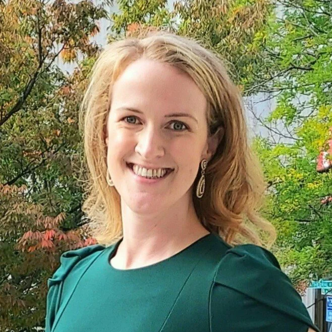 A woman with shoulder-length blonde hair smiling outdoors, wearing a teal dress and earrings, with colorful autumn trees in the background.