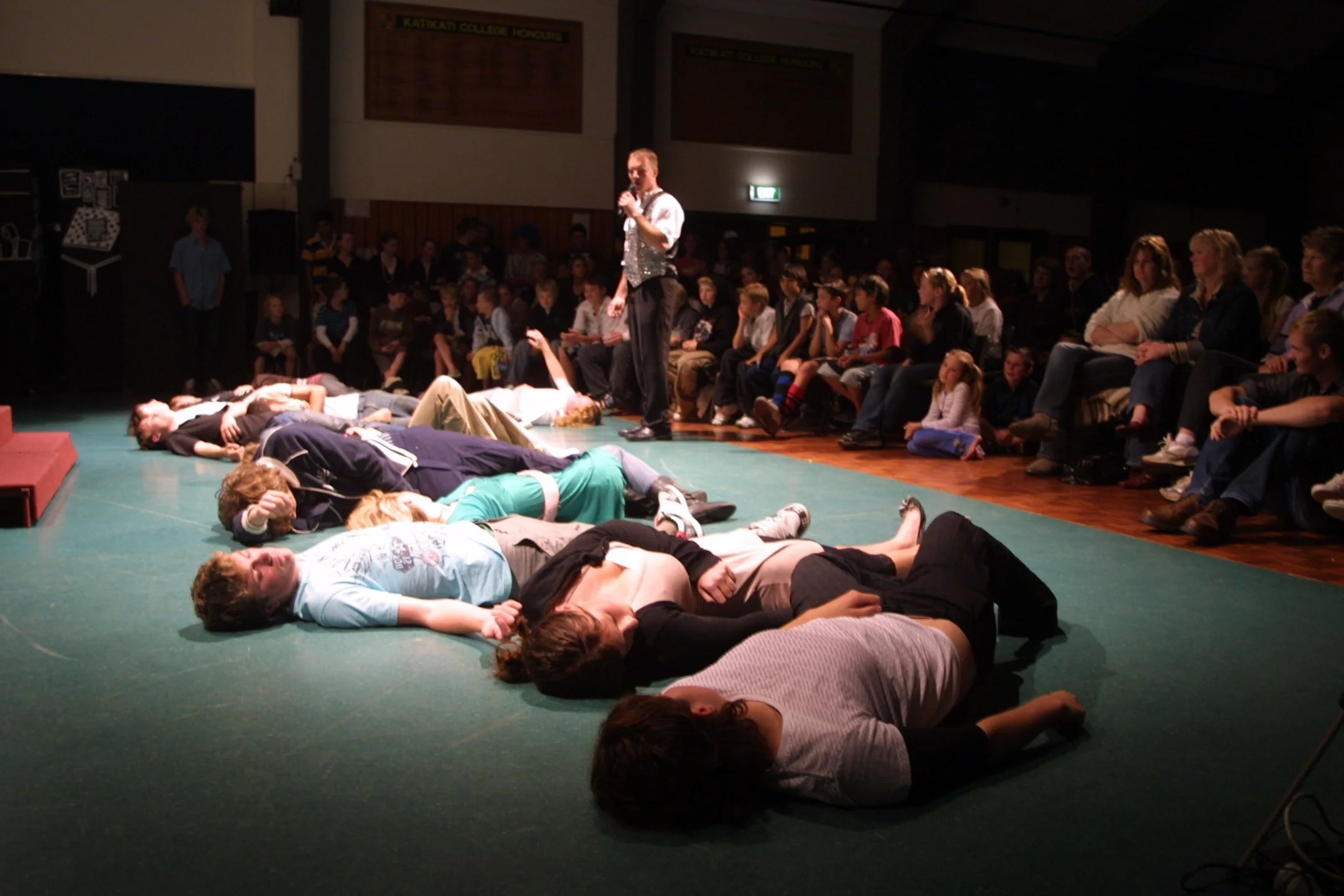 A group of people lying on the floor with their eyes closed, with a performer dressed in a vest standing nearby holding a microphone, hypnotist performance setting with an audience watching.