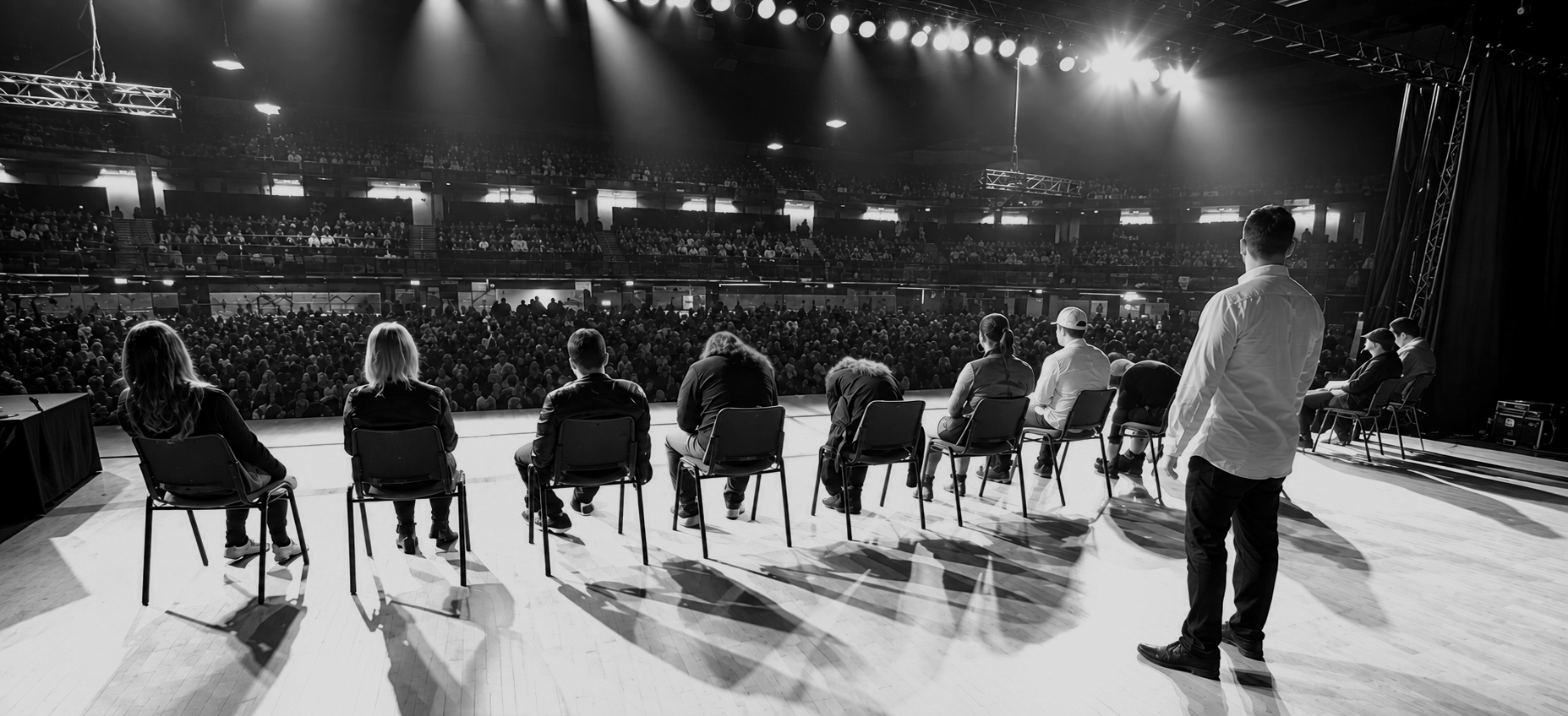 Black and white photo of a large auditorium filled with audience members watching a stage, with a panel of seven people seated facing the stage, and one person standing to the side.