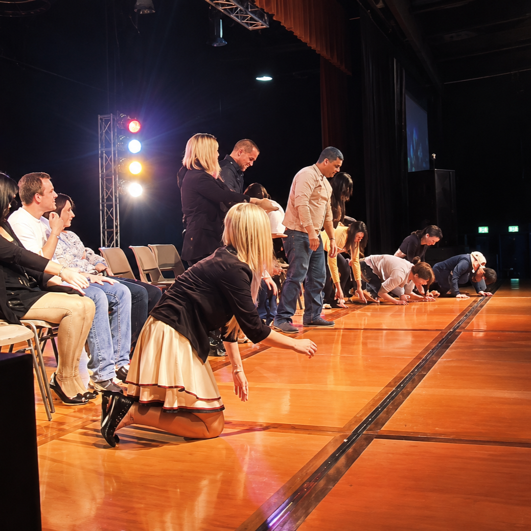 People seated and kneeling on a stage hypnotised, participating in a show with stage lights and a dark backdrop.