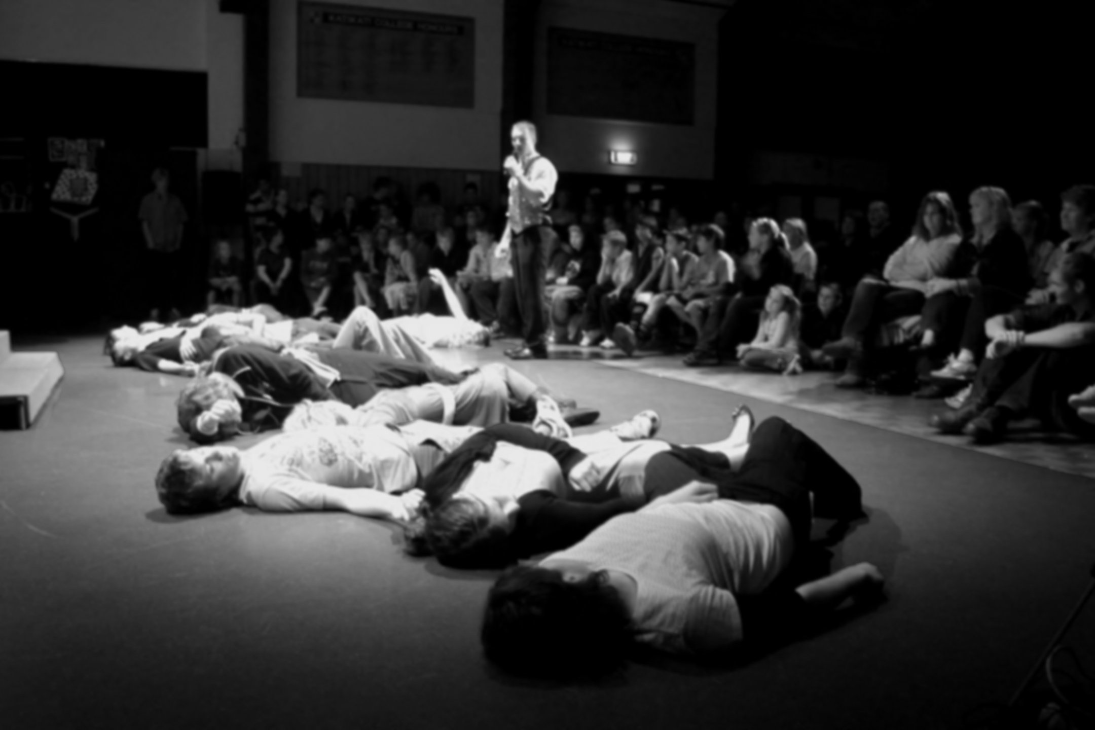 Black and white image of a person lying on the ground with their eyes closed, surrounded by several other people also lying down, while a man stands nearby speaking to an audience seated in a dark room.
