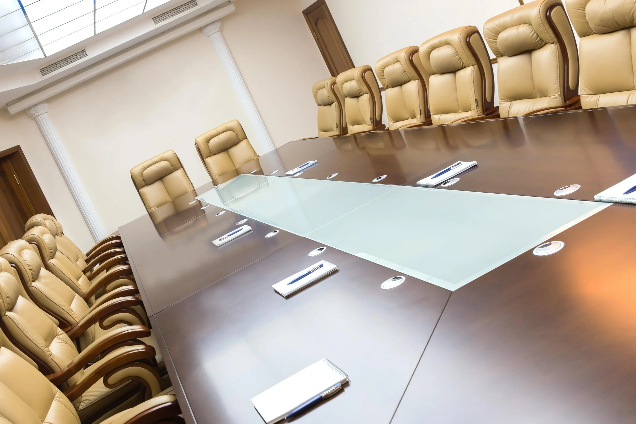 A conference room with a long wooden table surrounded by beige leather chairs, each equipped with pens and notepads.