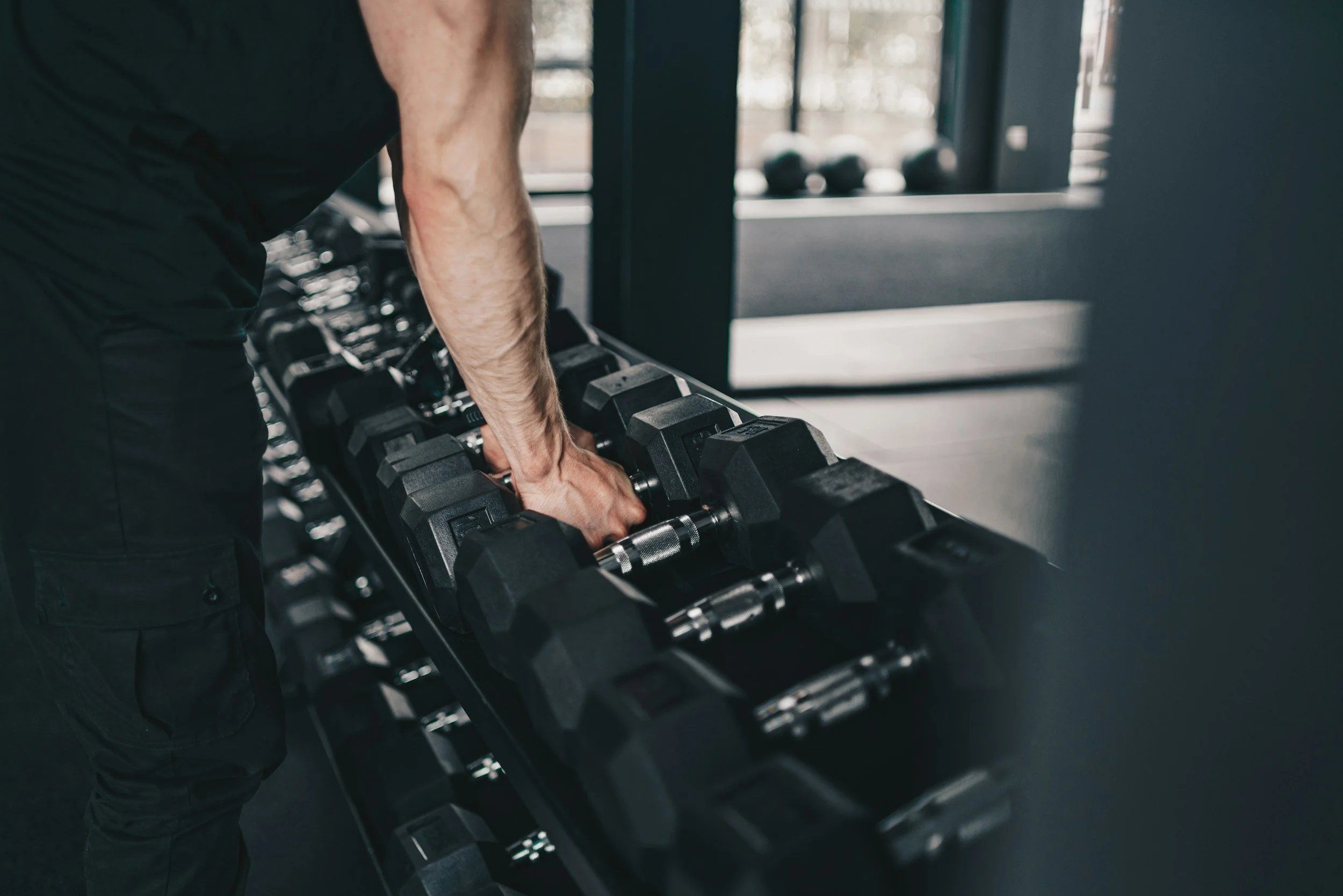 A person lifting black dumbbells in a gym, with weight racks and gym equipment visible in the background.