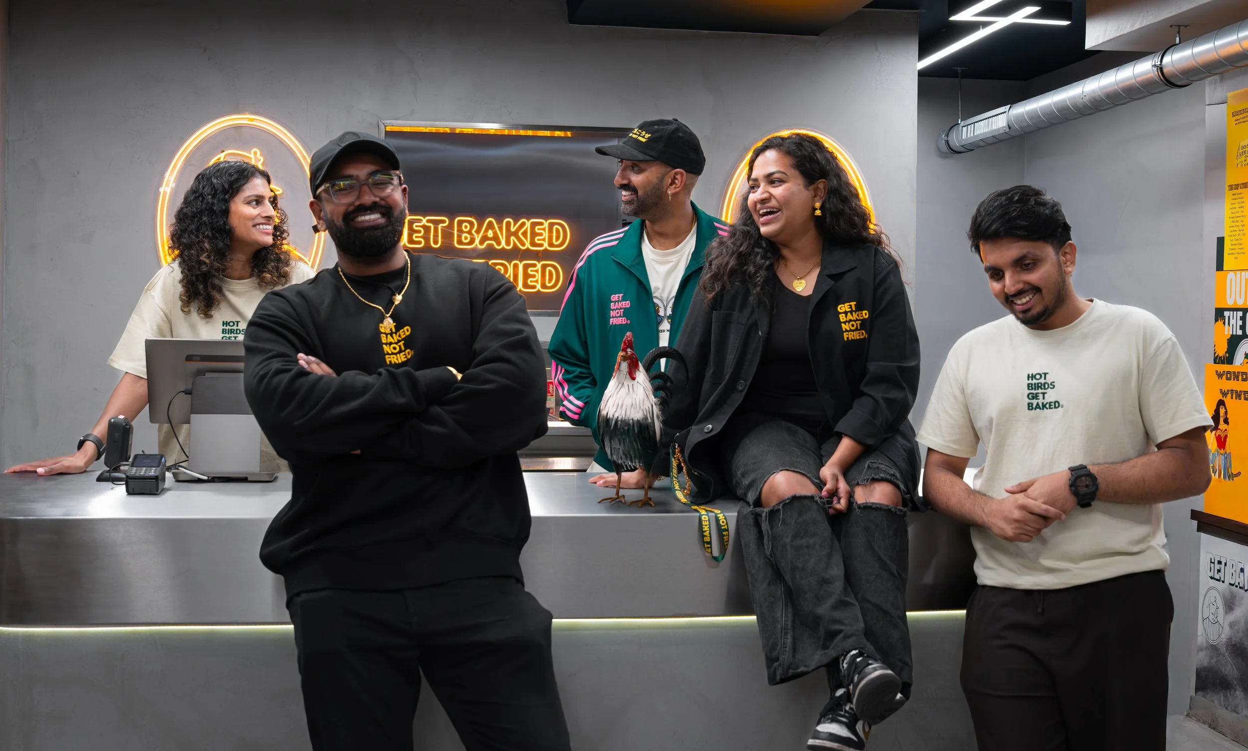 A group of five diverse people in a bakery or cafe, smiling and chatting. One woman is sitting on the counter with her legs crossed, a man standing with arms crossed, another woman standing beside him, and a woman and man on the right side. There is a neon sign that reads ‘Get Baked Not Fried’ behind them, with a rooster statue on the counter.