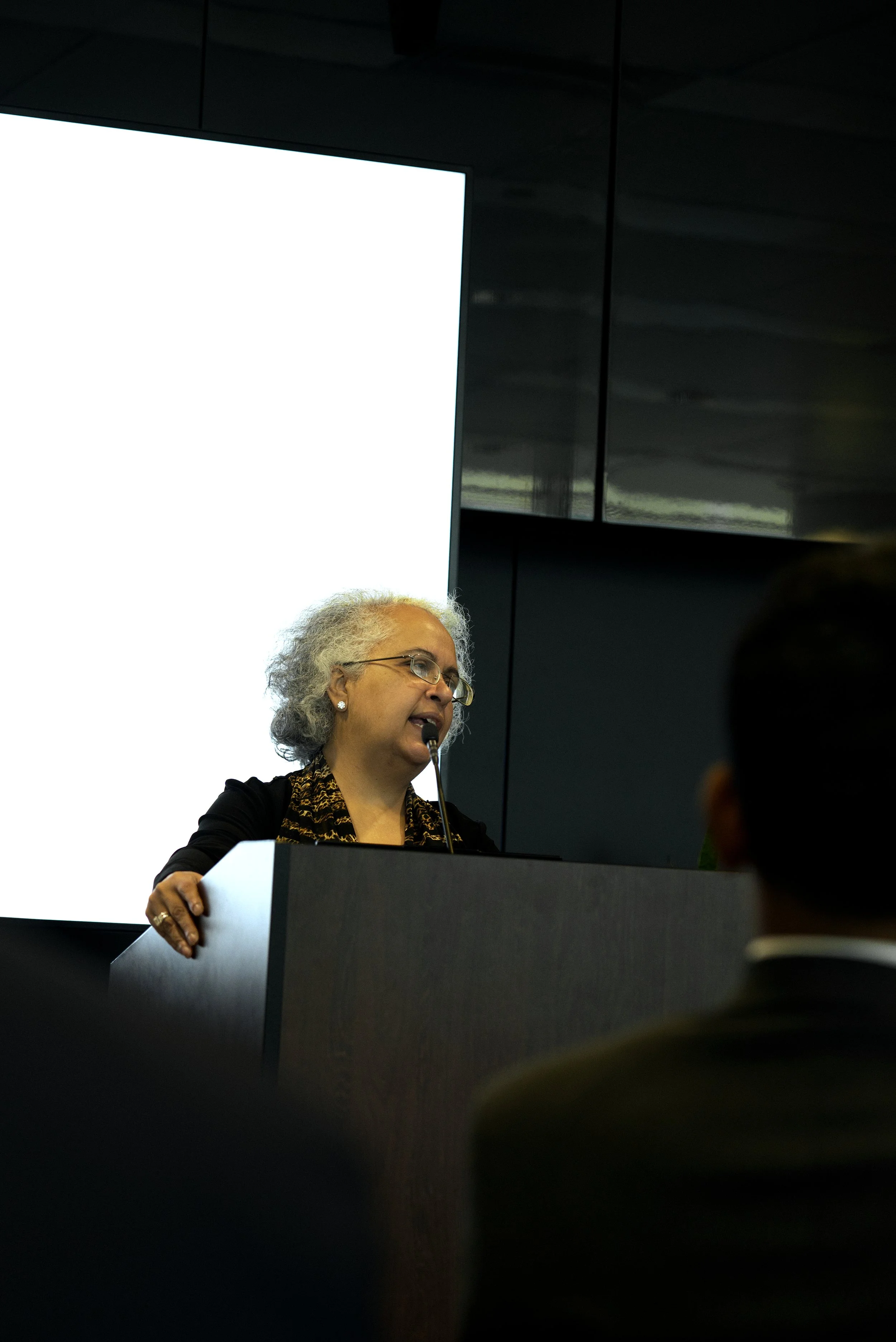 An older woman with curly gray hair and glasses speaking at a podium during a presentation or conference, with a large illuminated screen behind her.