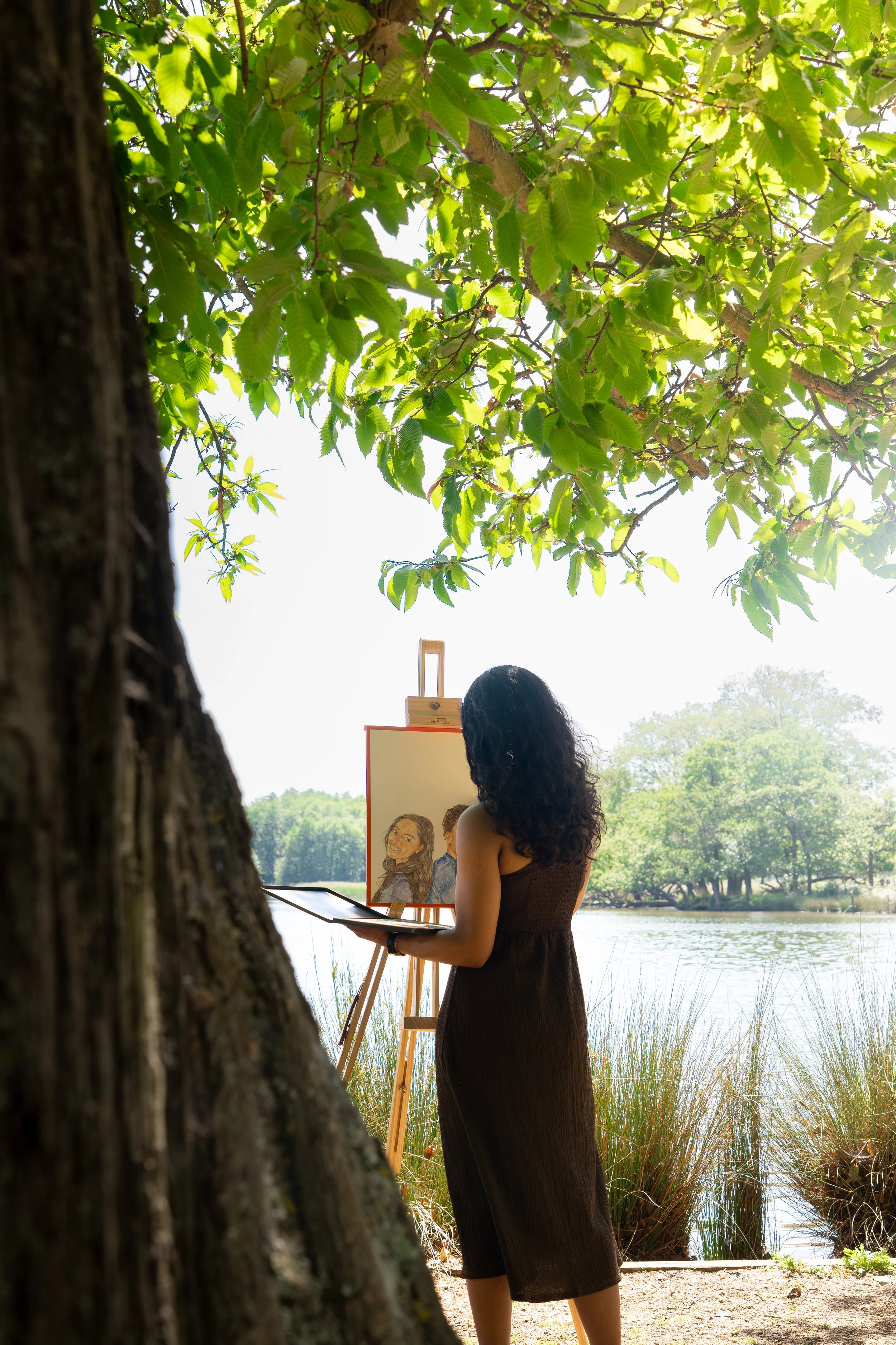A woman in a brown dress is painting a portrait outdoors near a lake, under the shade of trees with green leaves.