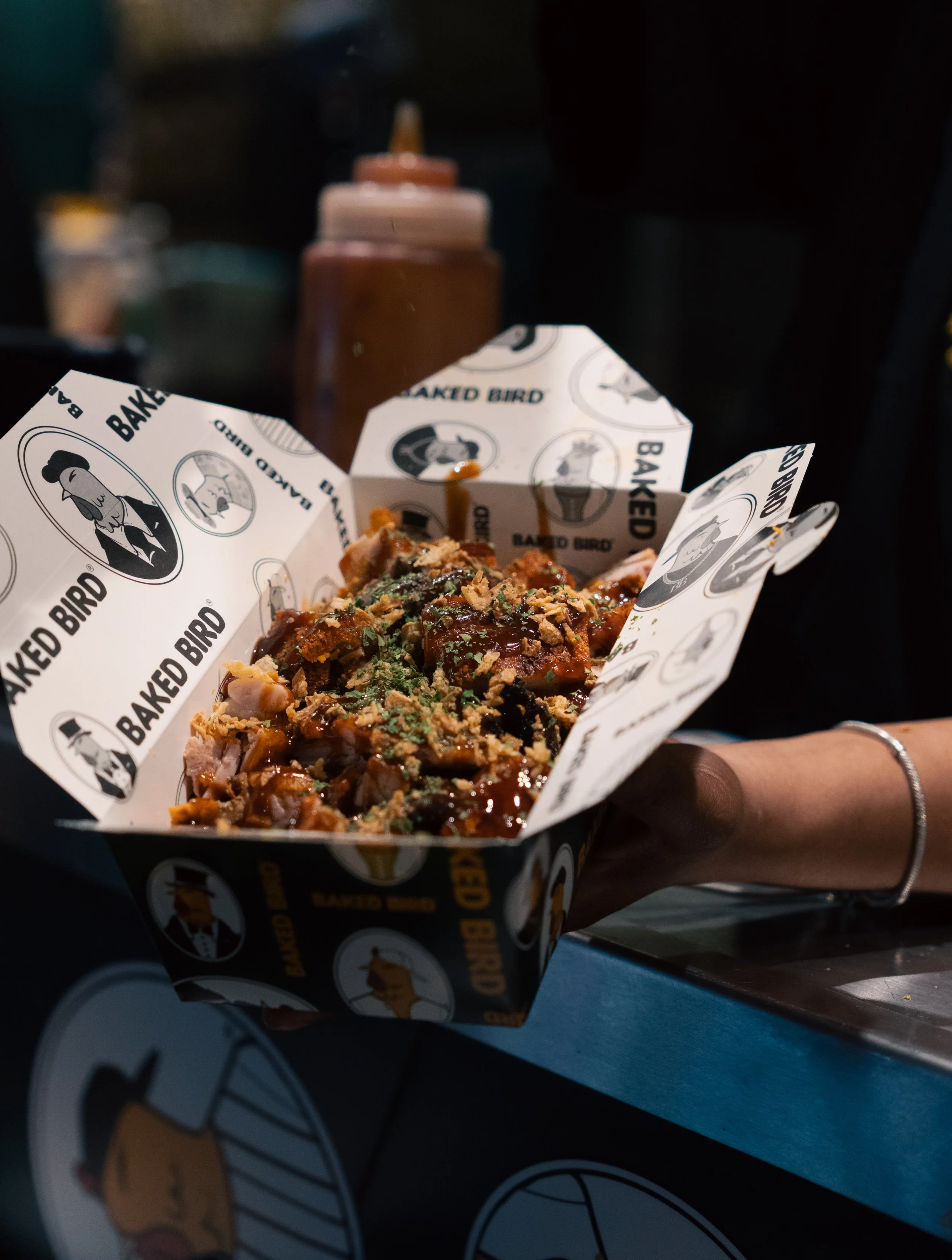 A hand holding a paper tray filled with Korean fried chicken topped with chopped green onions and herbs, with a bottle of sauce in the background, and a plate of fried chicken on a table.