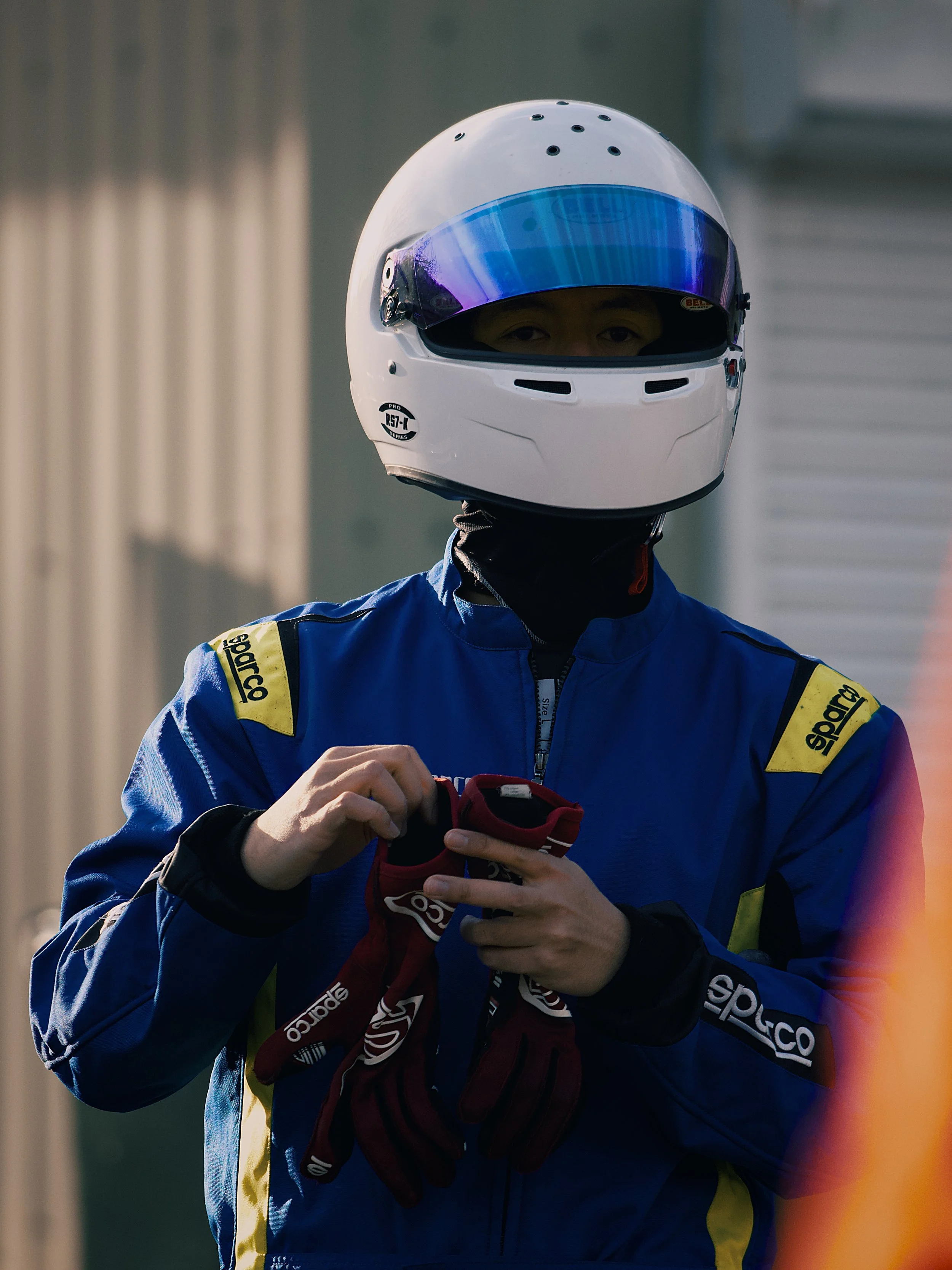 A race car driver wearing a white helmet with a blue visor and a blue racing suit with yellow and black accents, holding a red racing glove.