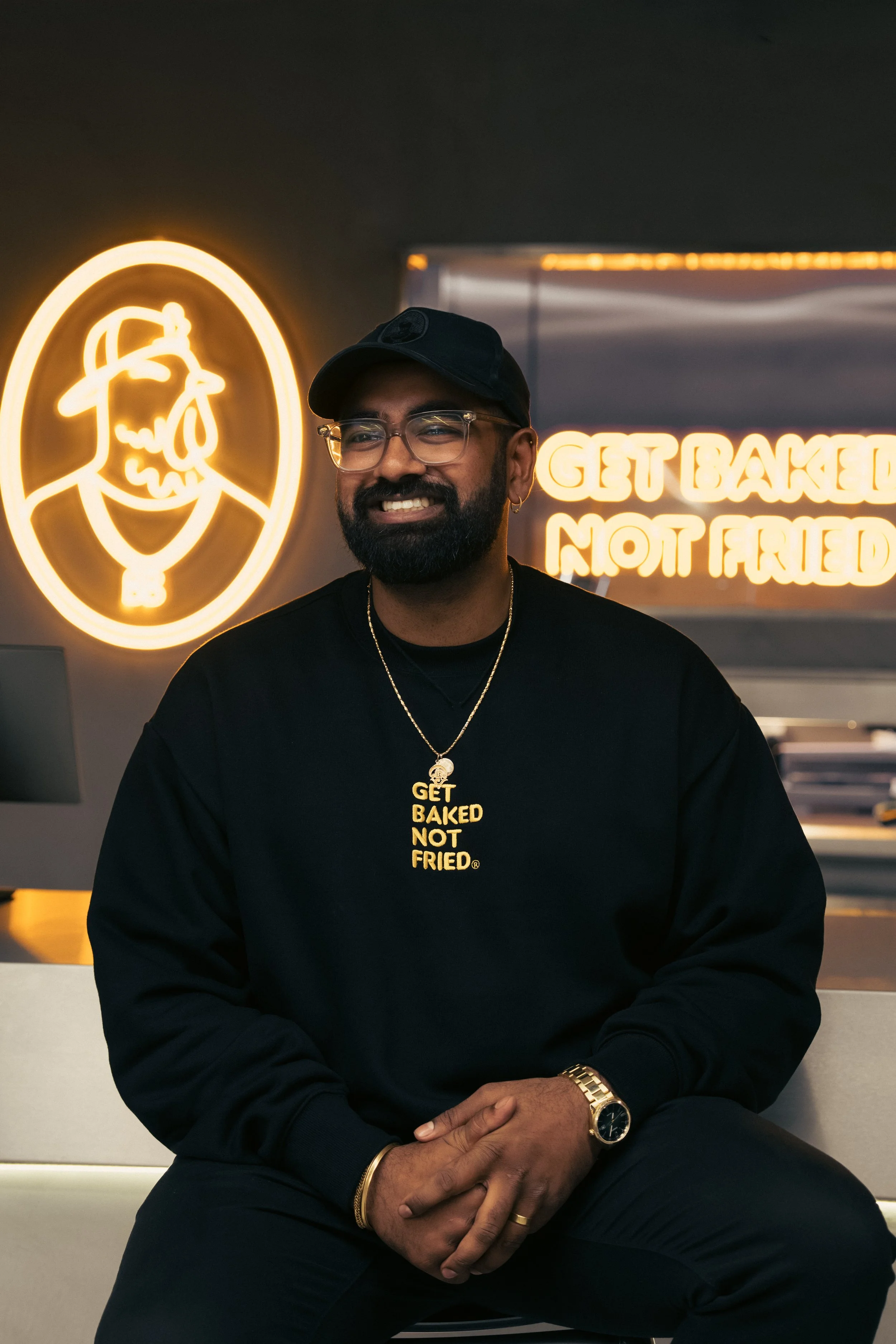 A man with glasses and a beard, wearing a black cap and black sweatshirt with yellow text, sitting with his hands clasped together. Behind him are neon signs, one with a cartoon chef face, and another with the text "GET BAKED NOT FRIED".