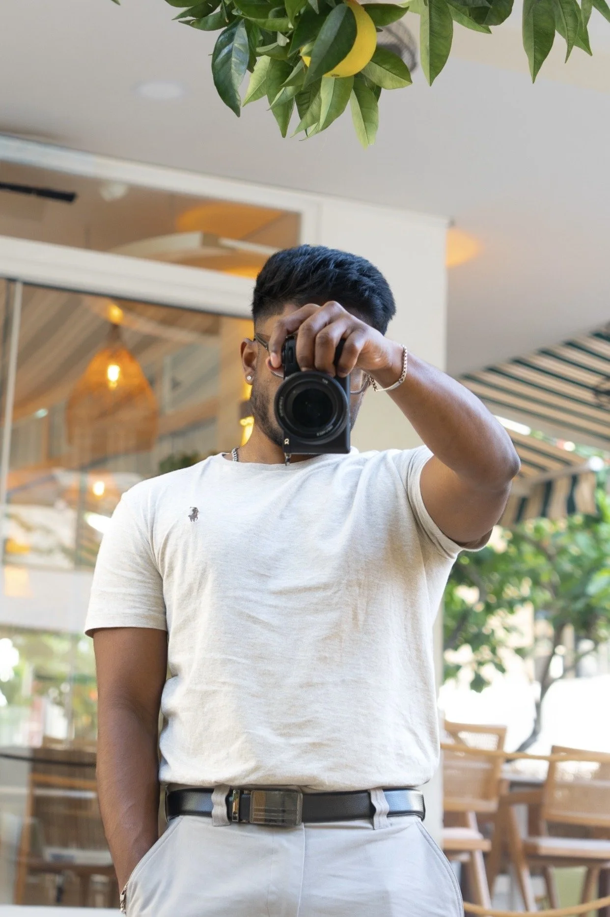 A person taking a selfie with a camera, standing outdoors in a cafe or restaurant patio with wooden furniture and greenery, wearing a light-colored T-shirt and grey pants.