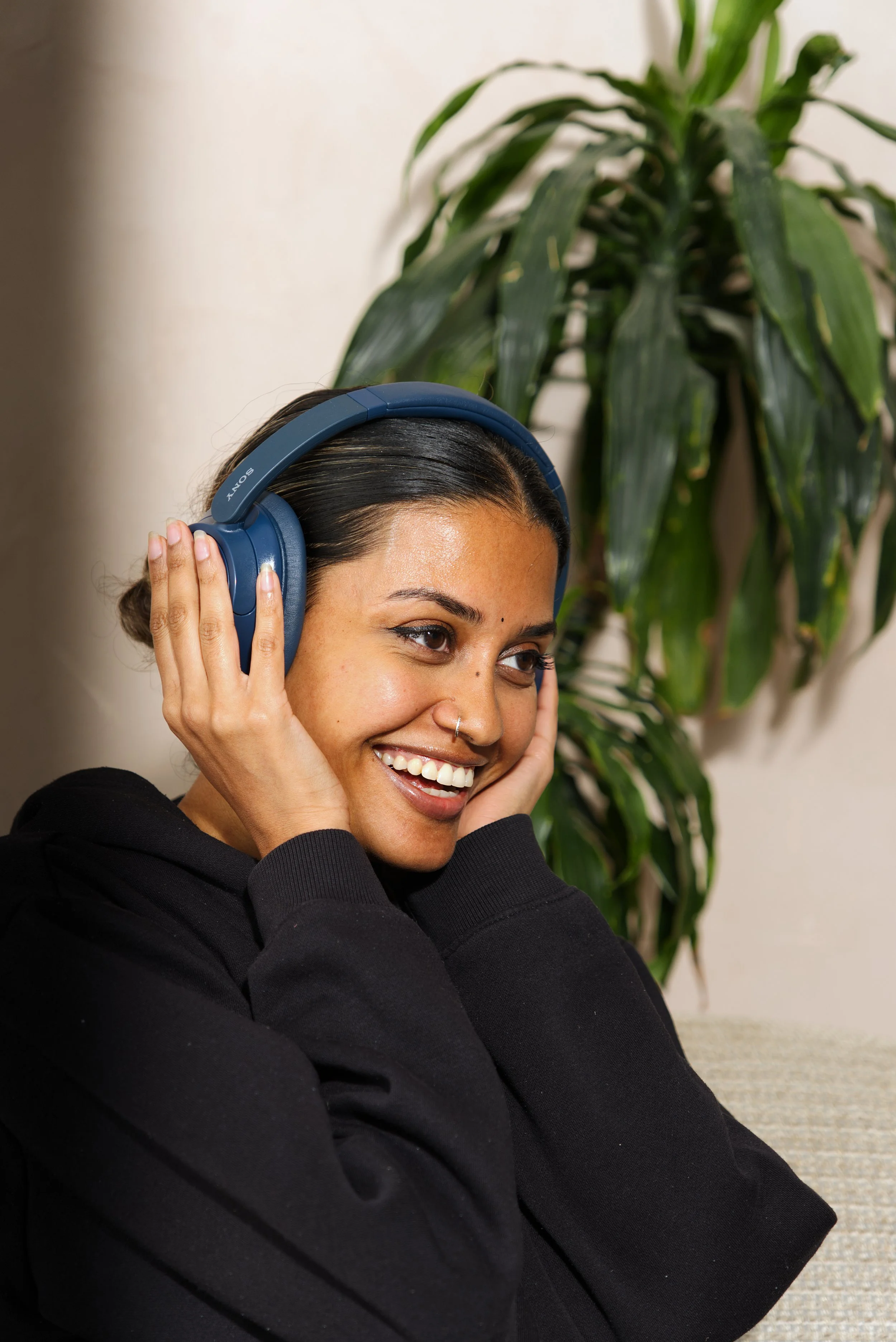A woman with dark hair and a nose piercing smiling while wearing blue over-ear Sony headphones, sitting indoors with a large plant with long green leaves in the background.