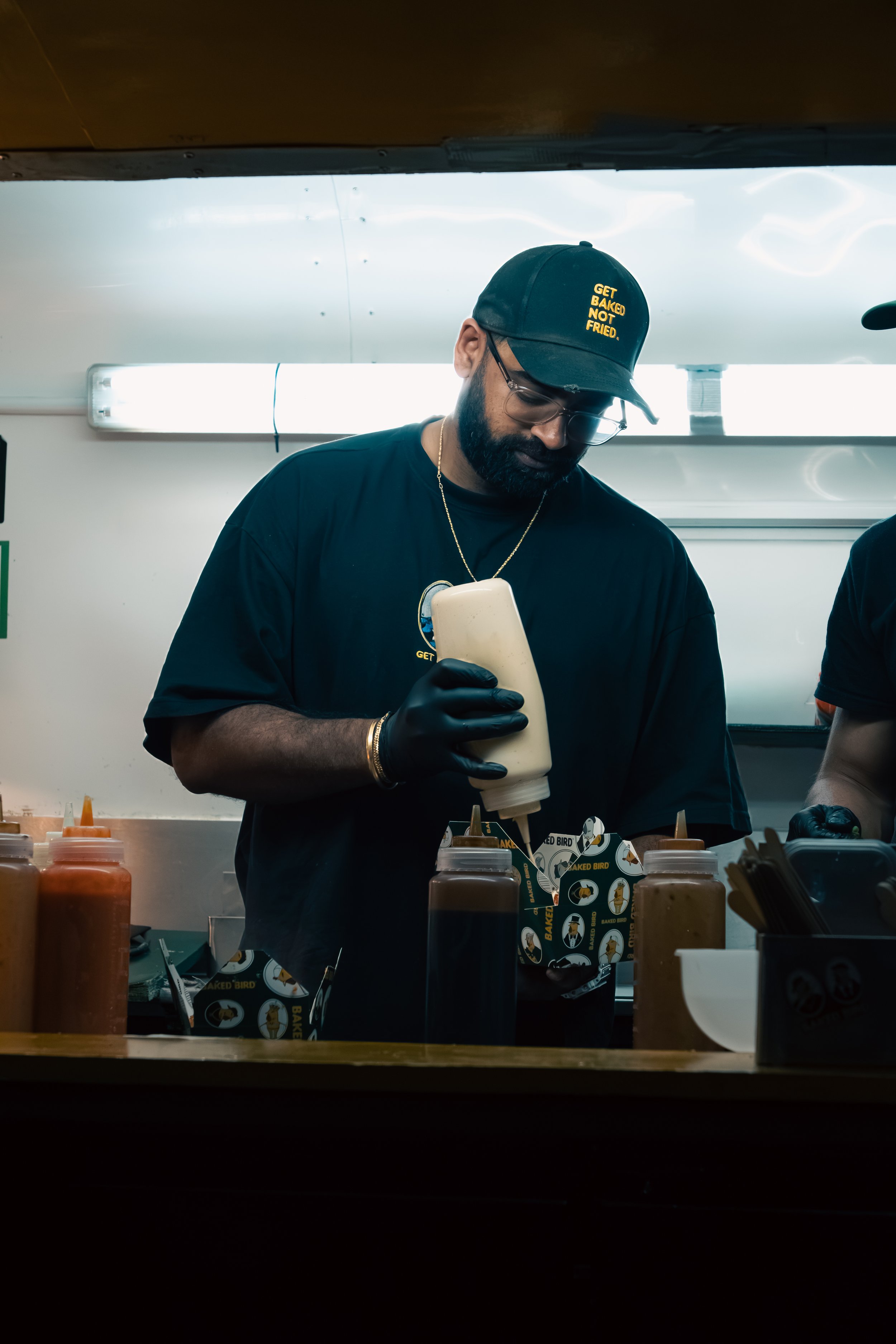 A man in a black shirt and glasses, wearing a black cap with yellow text, is pouring a sauce from a squeeze bottle over food. Several bottles of sauces and food containers are on the counter in front of him, and the background shows a kitchen or food stall environment.