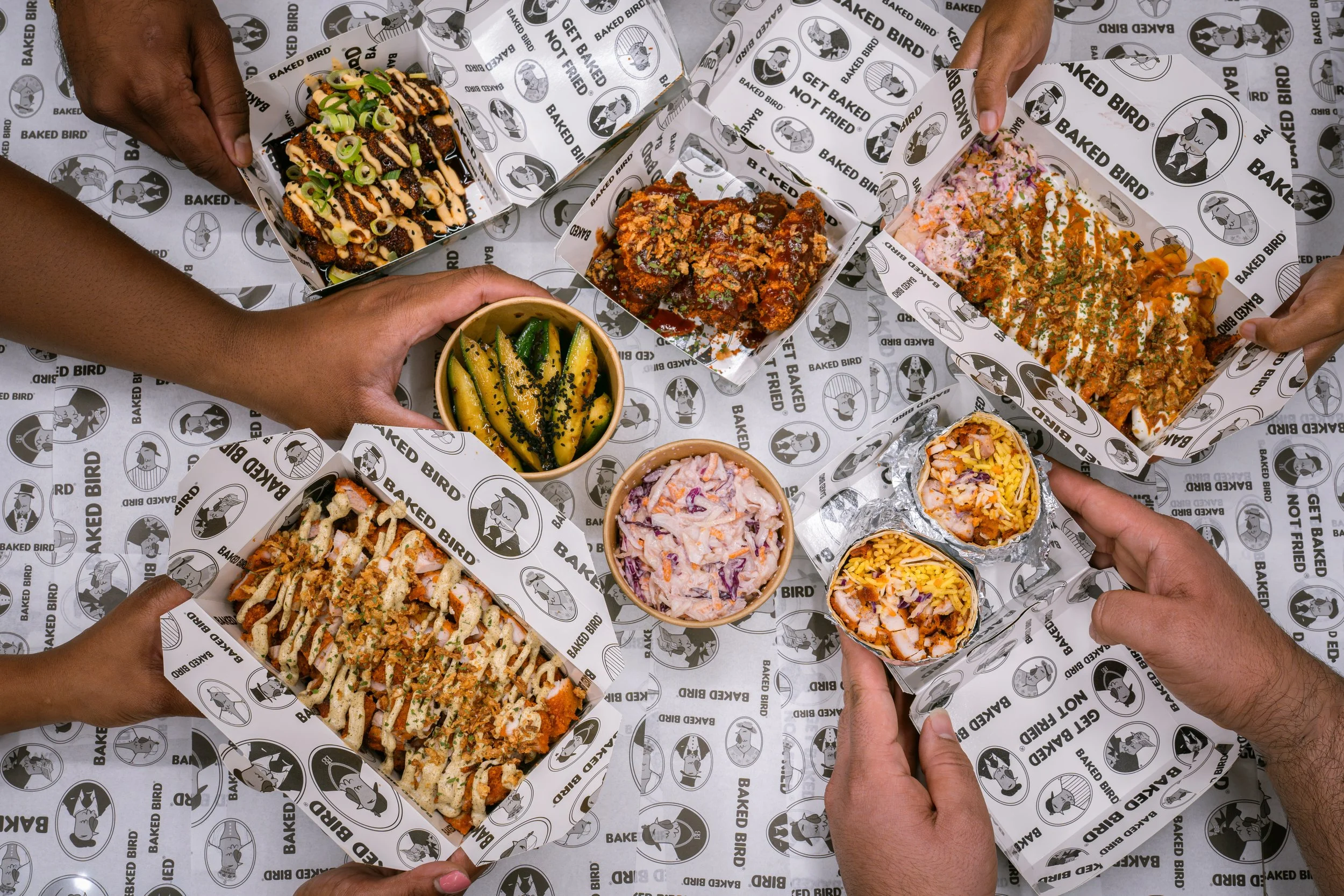 People holding boxes of baked food and small bowls of side dishes, with a patterned paper background.