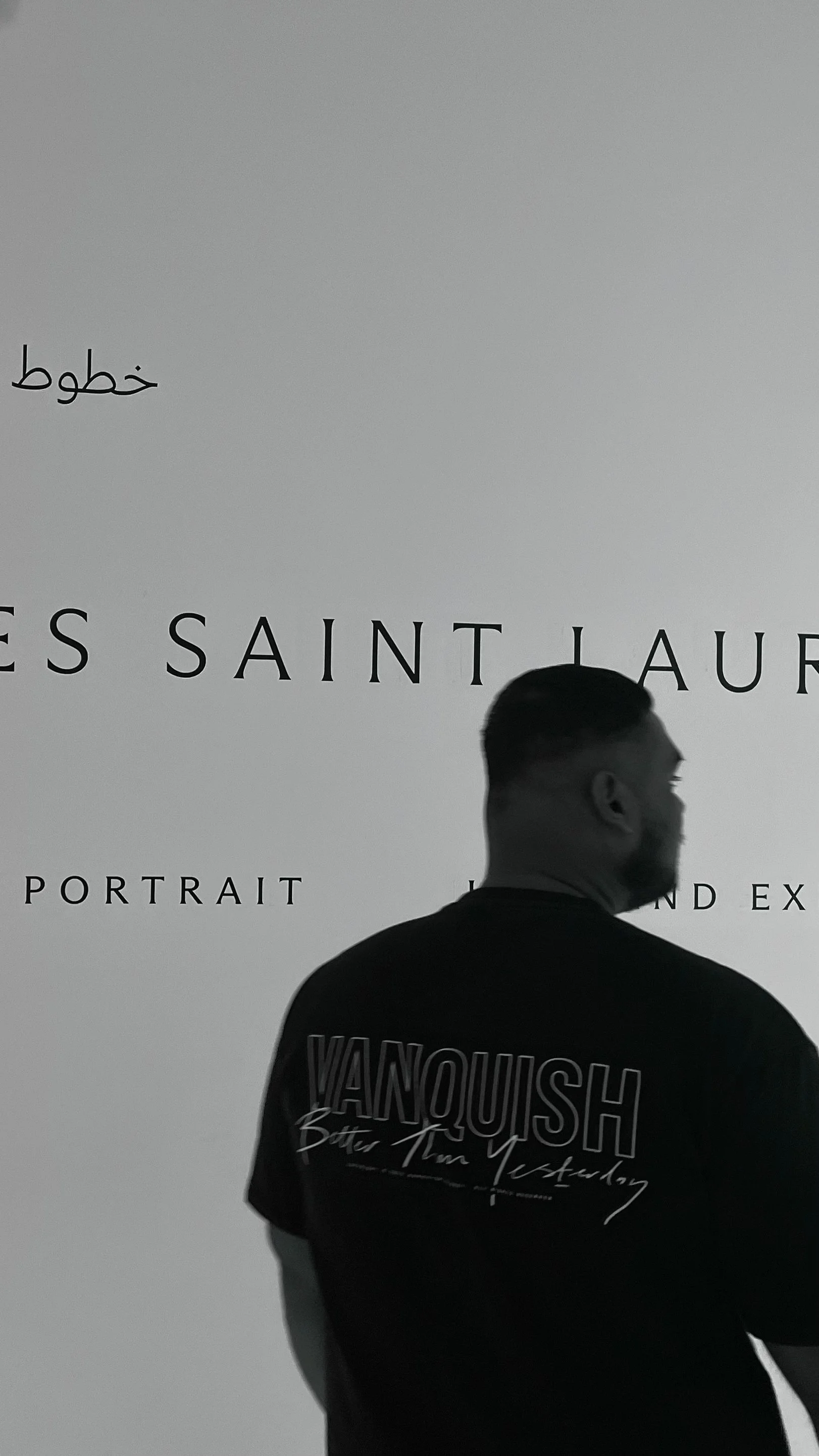 A man with a beard wearing a black t-shirt with the text "YANQUISH" on the back, standing in front of a white wall with black text and a small Arabic word.