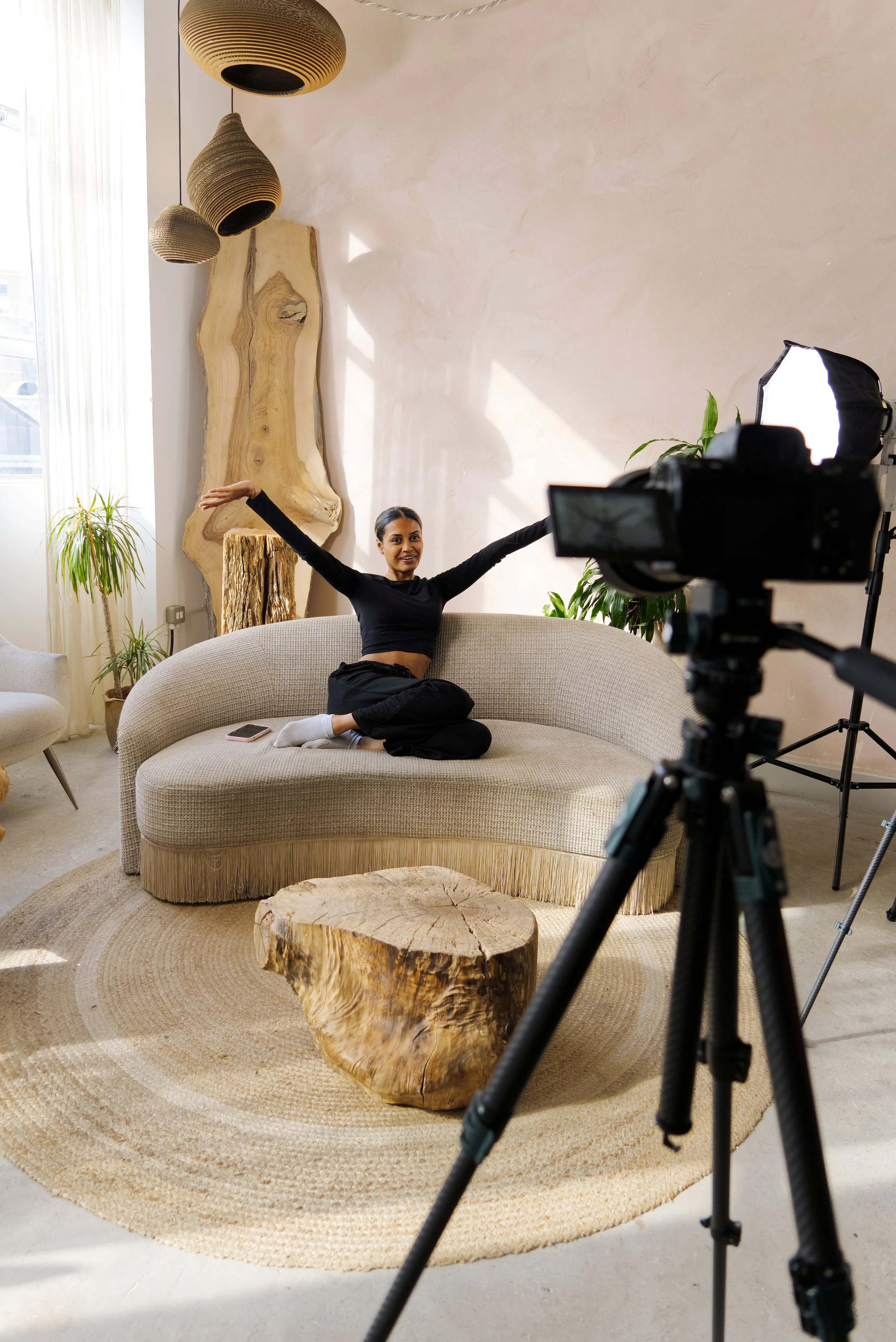 A woman sitting cross-legged on a beige sofa with her arms raised, being photographed in a well-lit living room with a large wooden wall art piece, plants, a round rug, and a camera set up on a tripod in front of her.