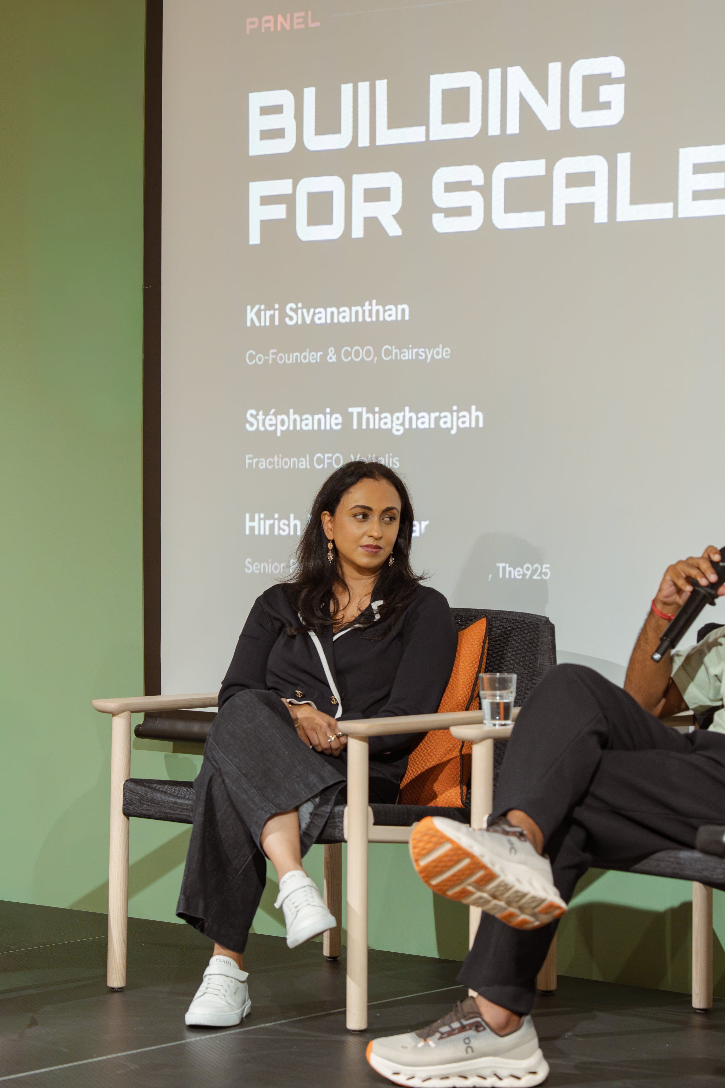 Two women are seated on a stage with a green background and a large screen behind them displaying a presentation slide titled "Building for Scale." The woman on the left has dark hair, is wearing a black blazer and gray pants, with her hands folded in her lap. She is looking slightly to her right with a neutral expression. The woman on the right is holding a microphone, wearing a light-colored shirt and dark pants, and appears to be speaking. There is a glass of water on a small table between them.