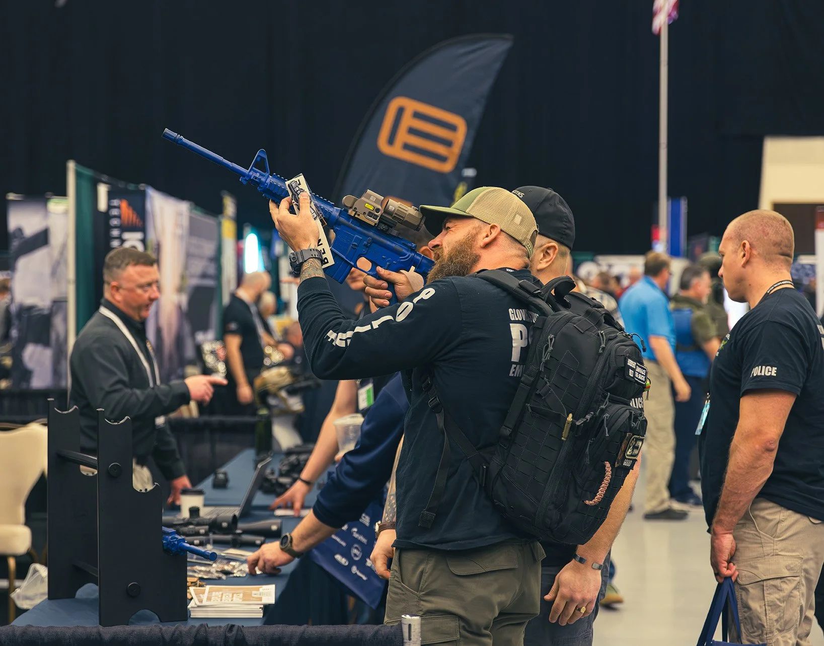 A man with a beard in police or tactical gear holding a blue rifle and aiming it at an exhibition booth at a trade show or convention.