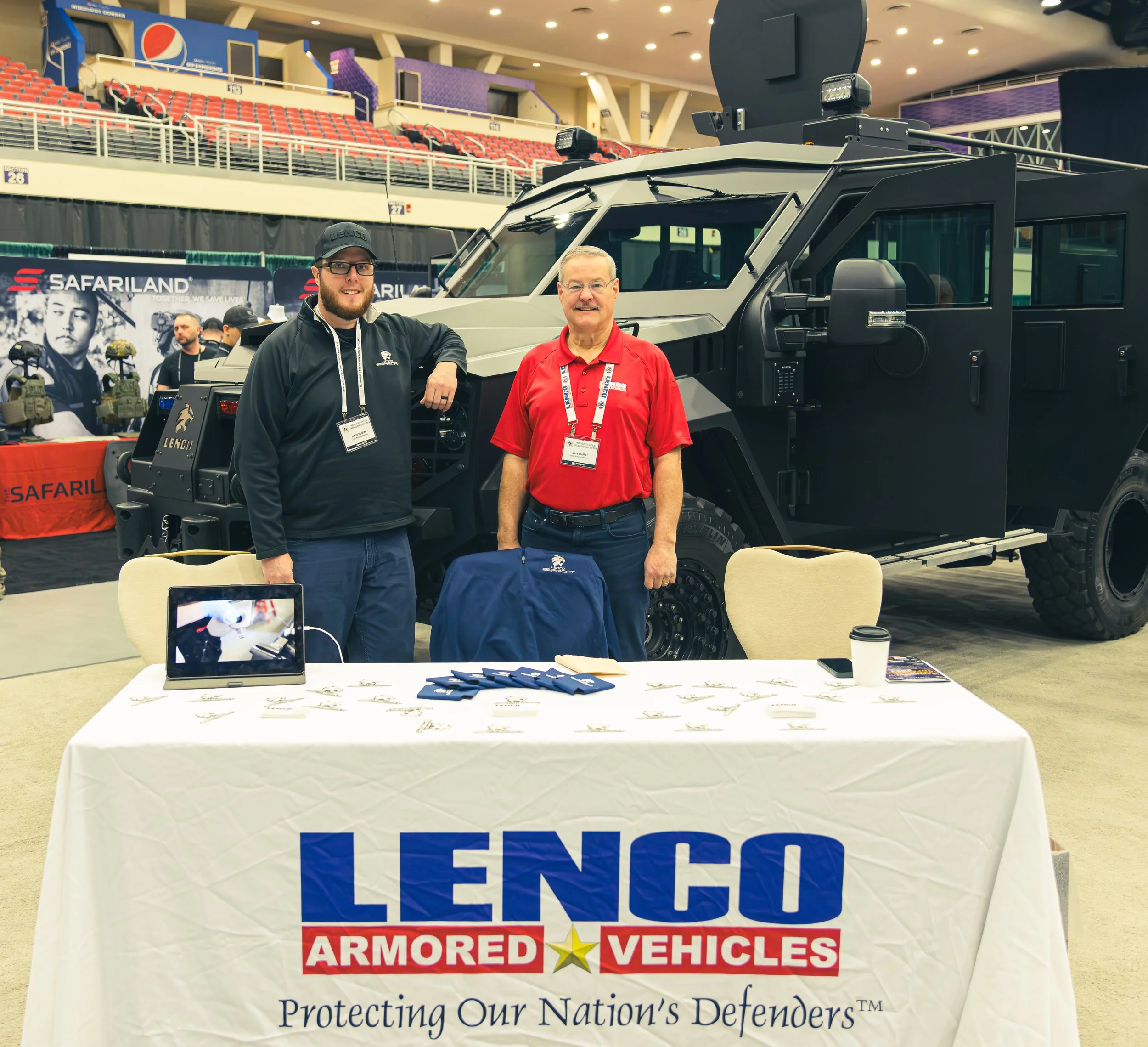Two men standing behind a table with a banner that reads "Lenco Armored Vehicles" and the tagline "Protecting Our Nation's Defenders." They are at an indoor event with an armored vehicle in the background. The man on the left is wearing a black jacket and cap, and the man on the right is wearing a red polo shirt with a lanyard and badge.
