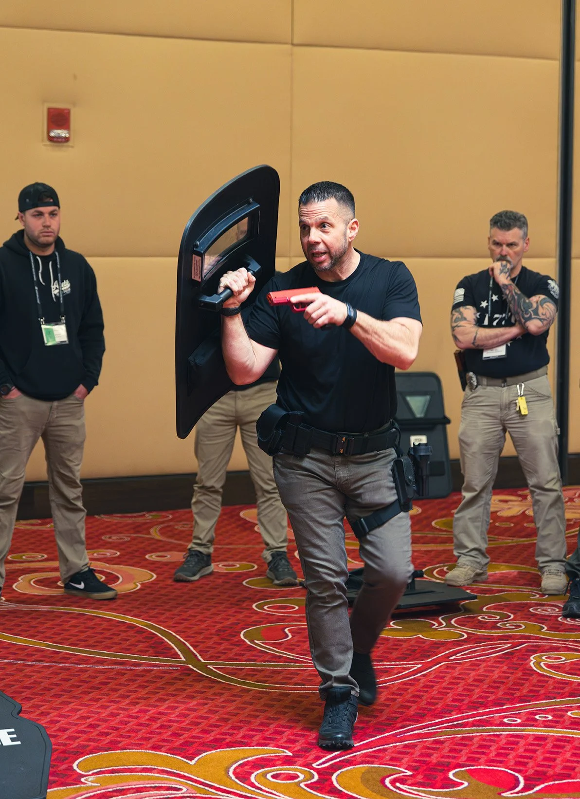 Man holding a shield and a gun, leading a training session for three men with tattoos and casual clothing, in a conference room with red patterned carpet.