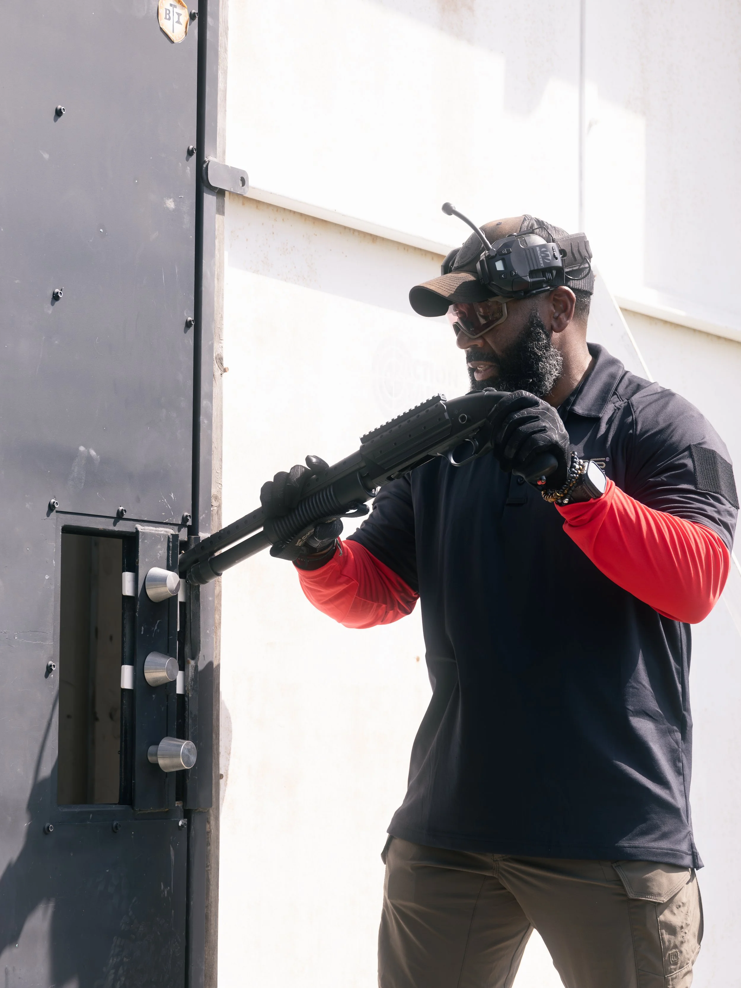 A man wearing sunglasses, a baseball cap, gloves, and tactical gear is holding a shotgun and aiming it at a door. He is standing outside near the door.