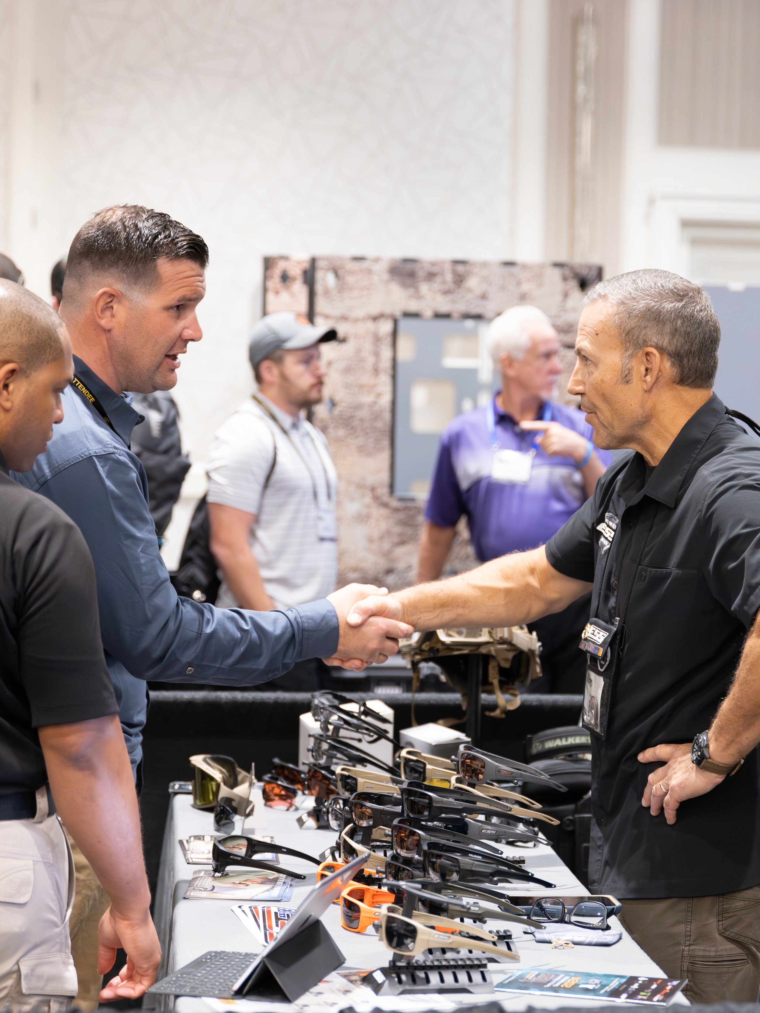 People shaking hands at a table displaying sunglasses and eyewear on a busy indoor event or trade show.