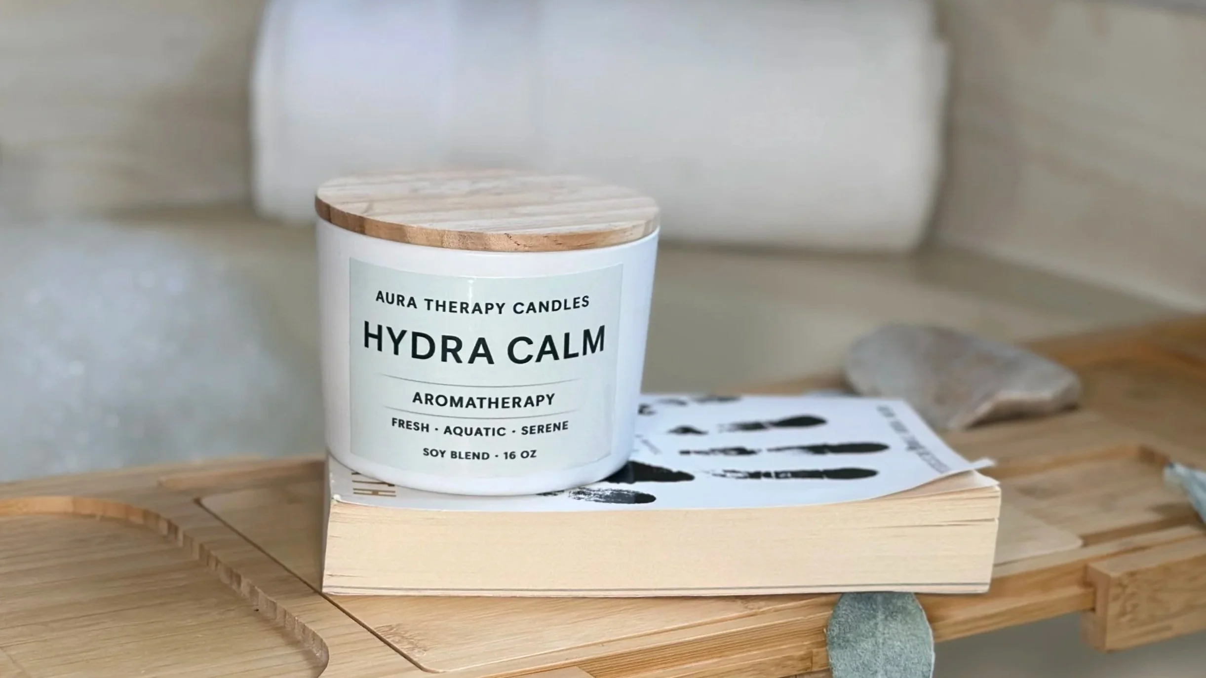A white container with a wooden lid labeled 'Aura Therapy Candles, Hydra Calm, Aromatherapy, Fresh, Aquatic, Serene, Soy Blend, 16 oz' placed on a wooden table.