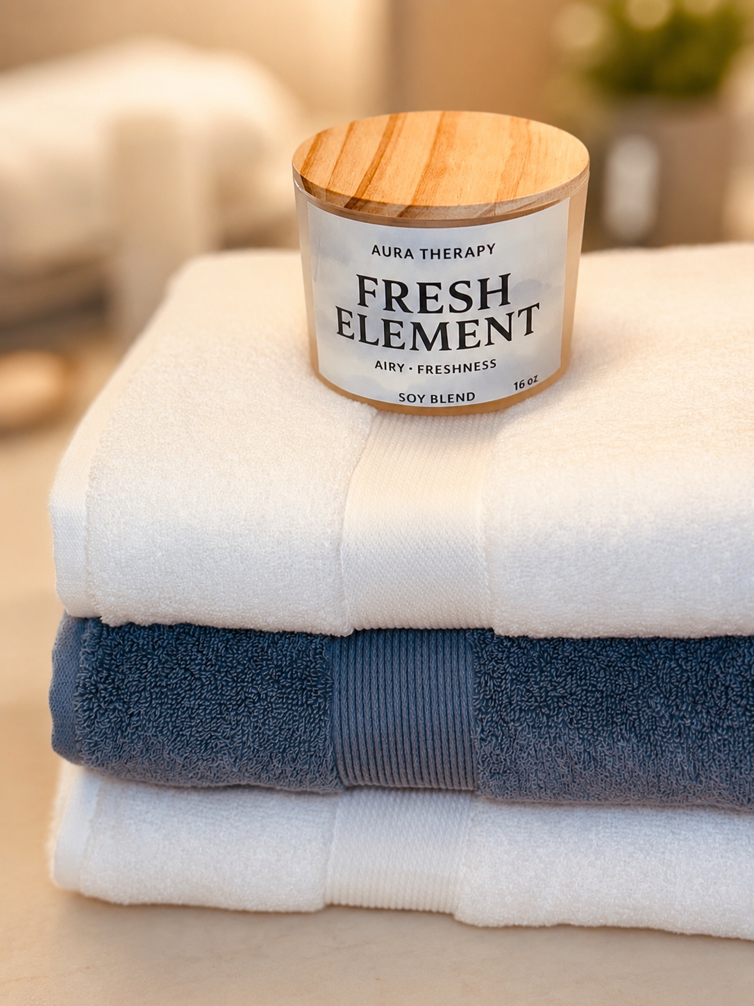 Stacked white and gray towels with a wooden lid candle labeled 'Fresh Element Aura Therapy Soy Blend, Airy Freshness, 16 oz' on top.